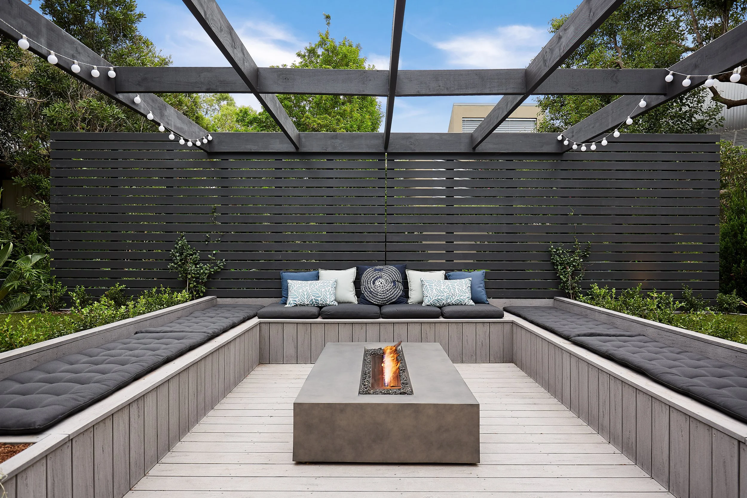 Outdoor patio with U-shaped wooden bench seating, gray cushions, decorative pillows, a concrete fire pit in the center, and string lights overhead, enclosed by a wooden privacy screen with greenery behind.