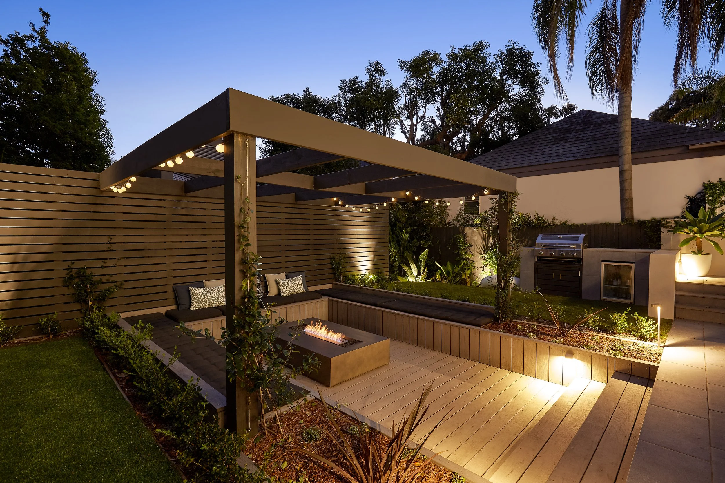 An outdoor backyard patio at dusk with a wooden pergola, a fire pit, built-in bench seating with cushions, a gas grill, and garden lighting, surrounded by a fence and lush greenery.