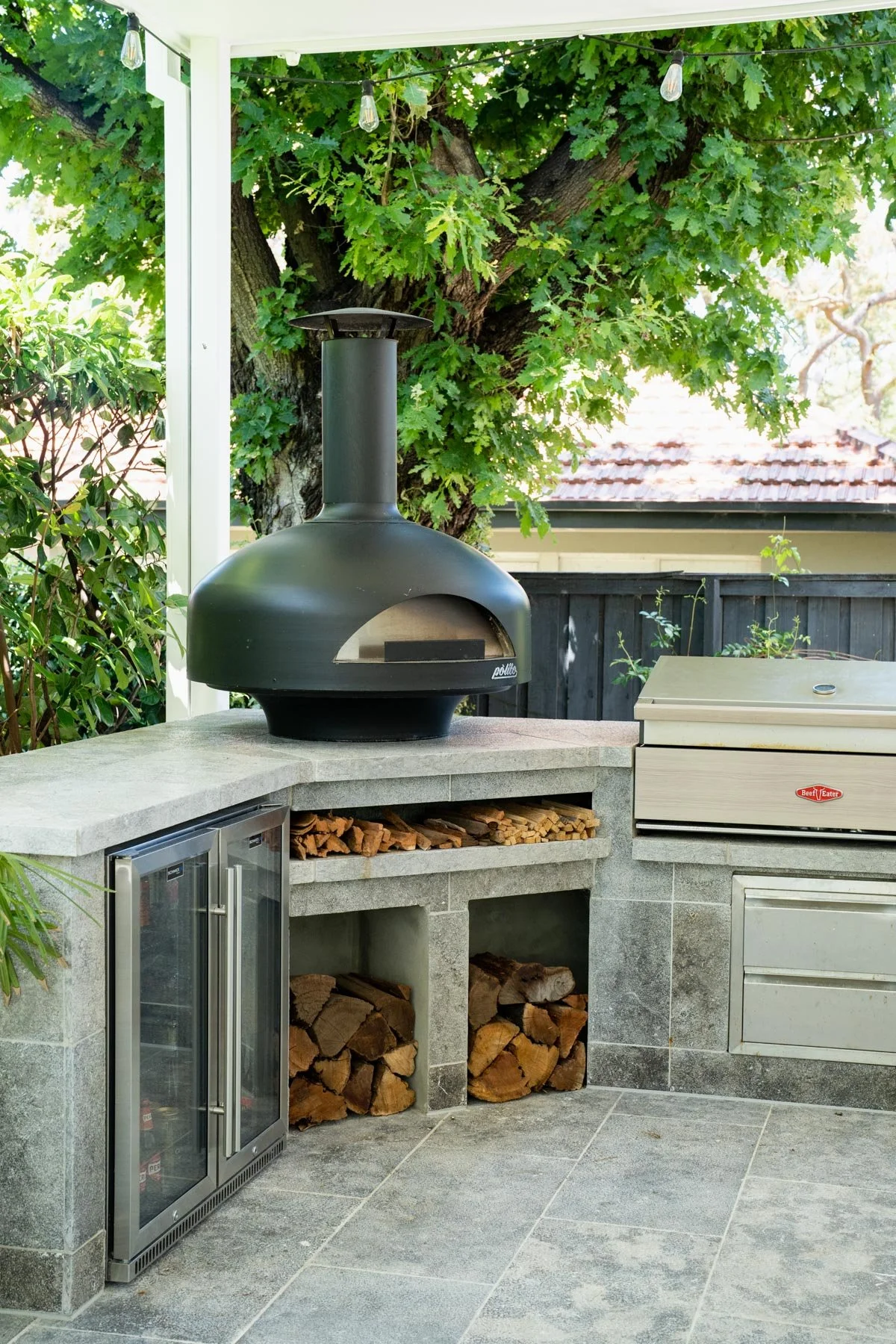 Outdoor kitchen with a black pizza oven, stacked firewood beneath, a small refrigerator, and a grill, set against greenery and a large tree.