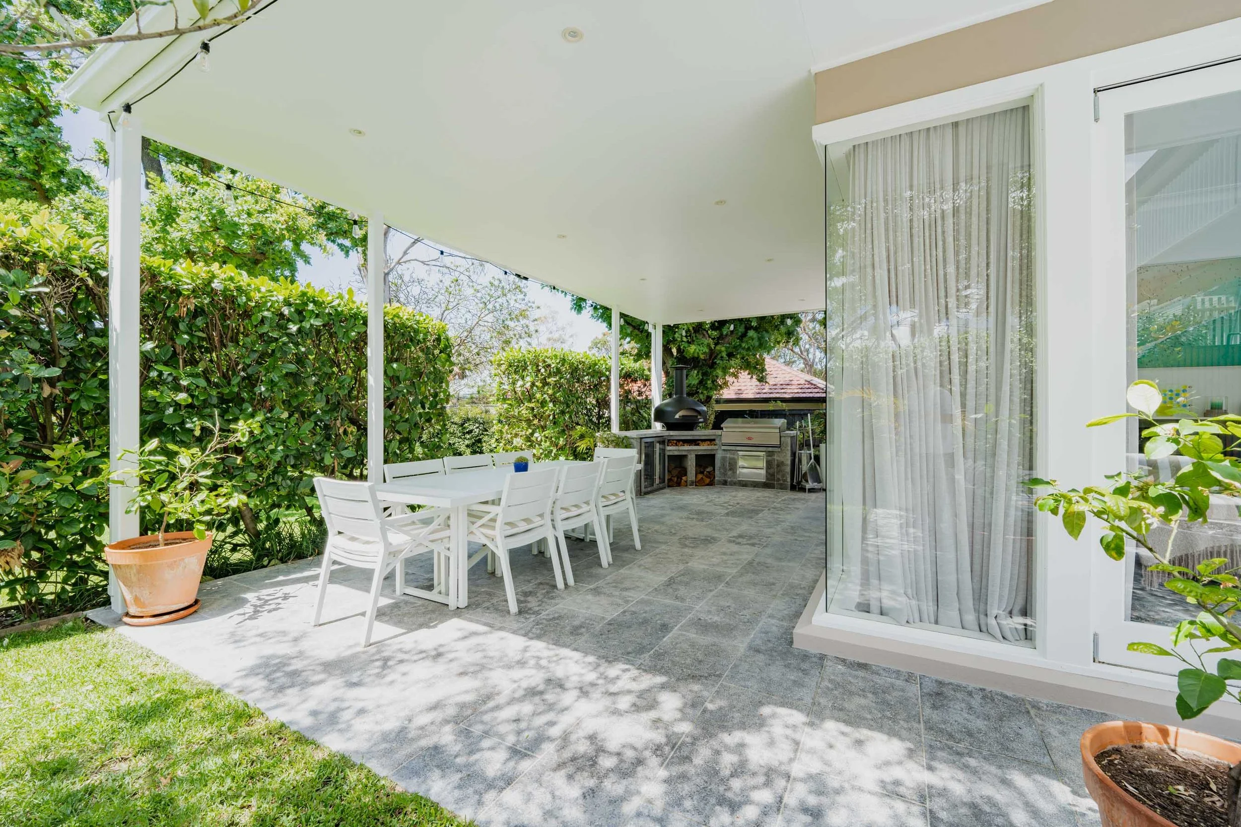 Outdoor patio area with a white dining table and chairs, a barbecue grill, a potted plant, and a sliding glass door with curtains.