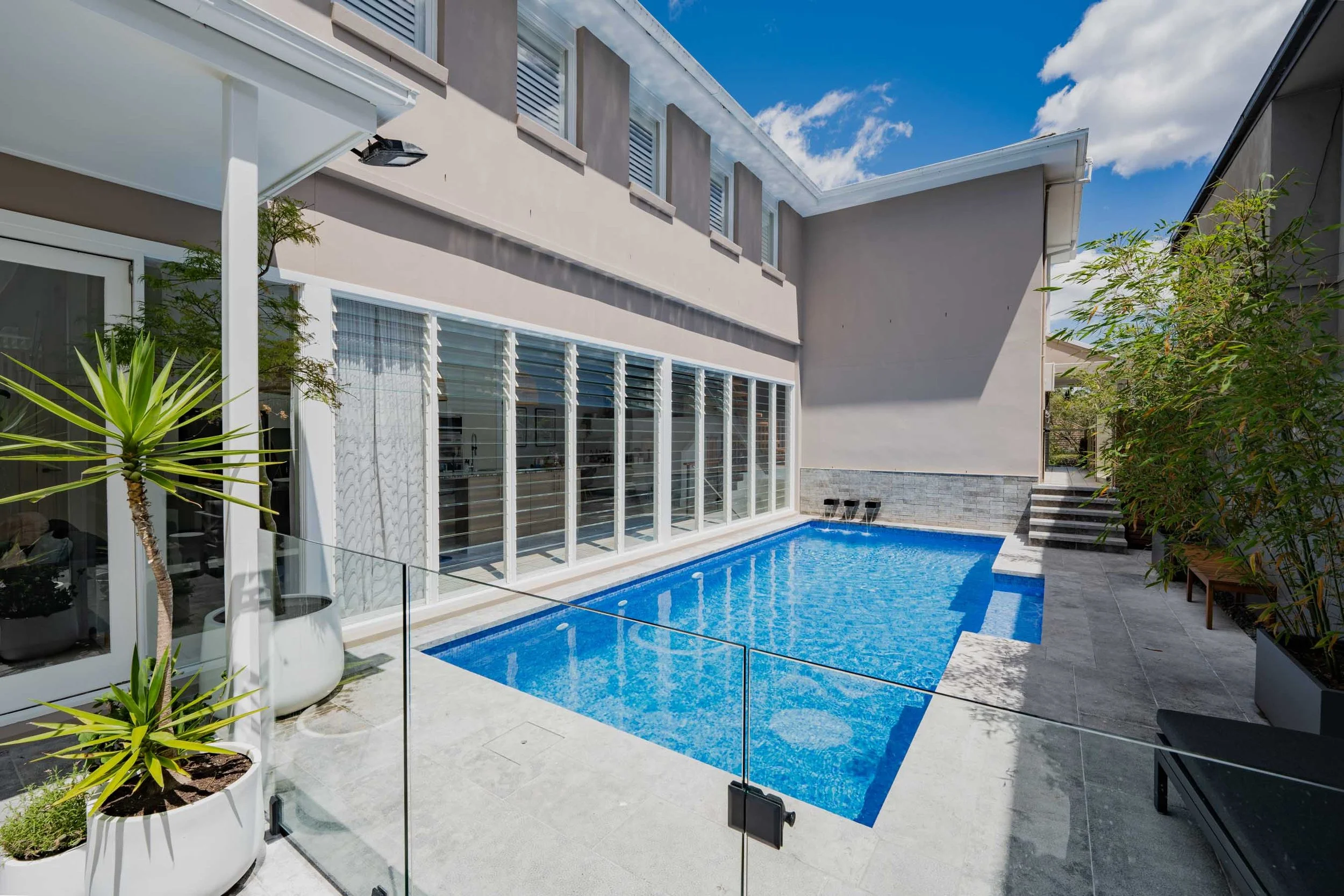 Modern backyard with a small rectangular swimming pool, potted plants, and a glass fence.