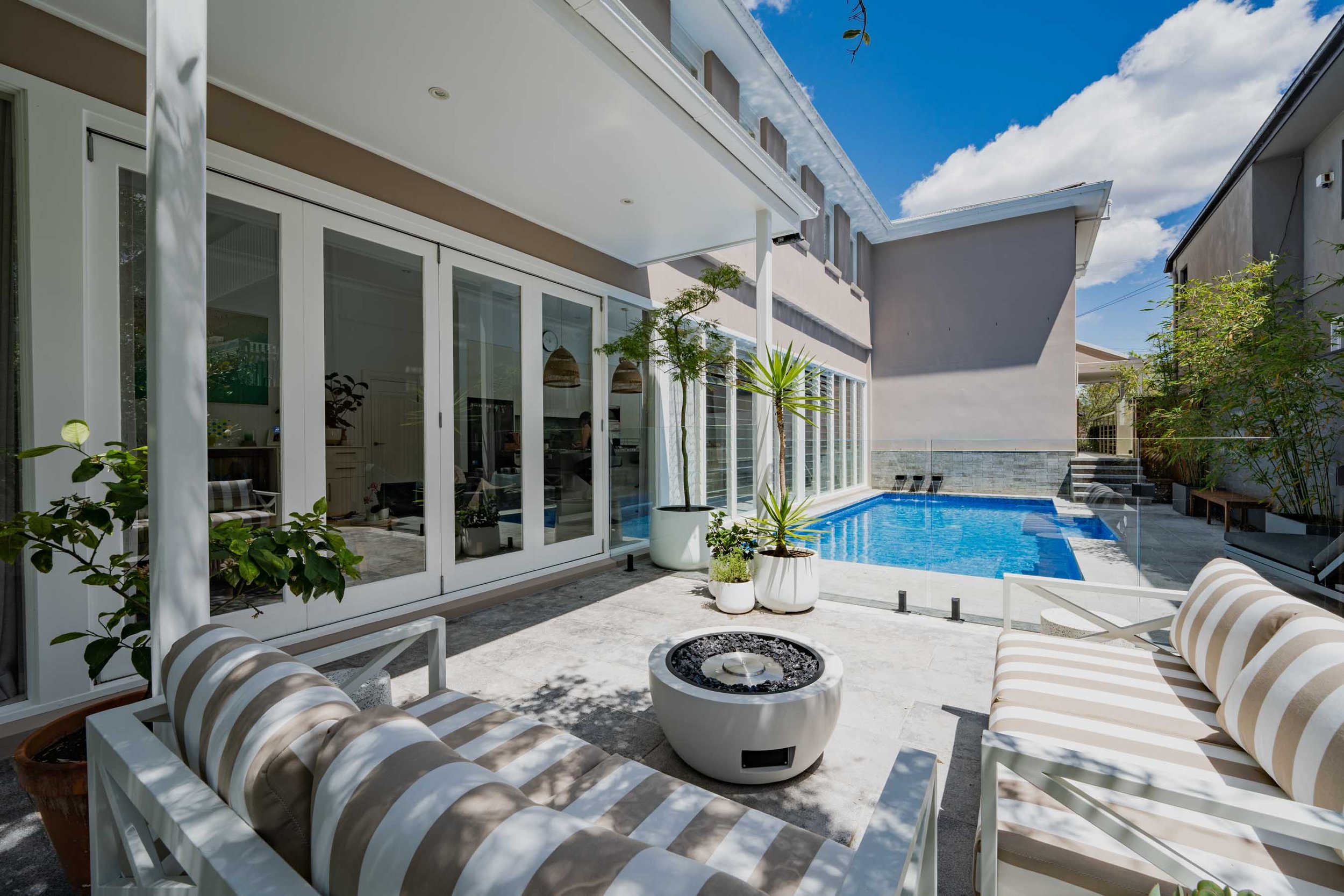 Modern backyard with a swimming pool, outdoor seating with striped cushions, potted plants, and a glass-enclosed house with sliding doors under a partly cloudy sky.