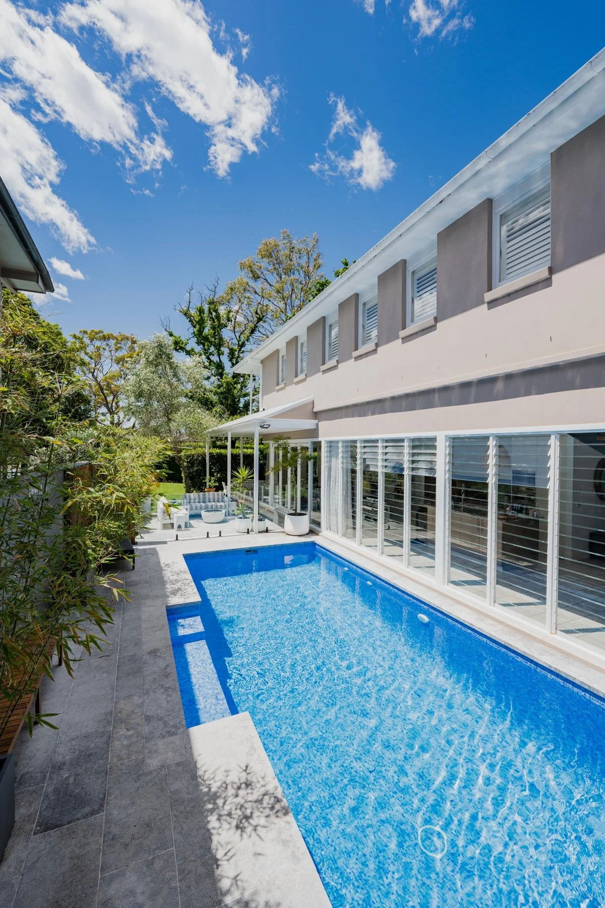 A backyard with a swimming pool next to a modern house with large windows, surrounded by greenery and tall trees under a clear blue sky with few clouds.