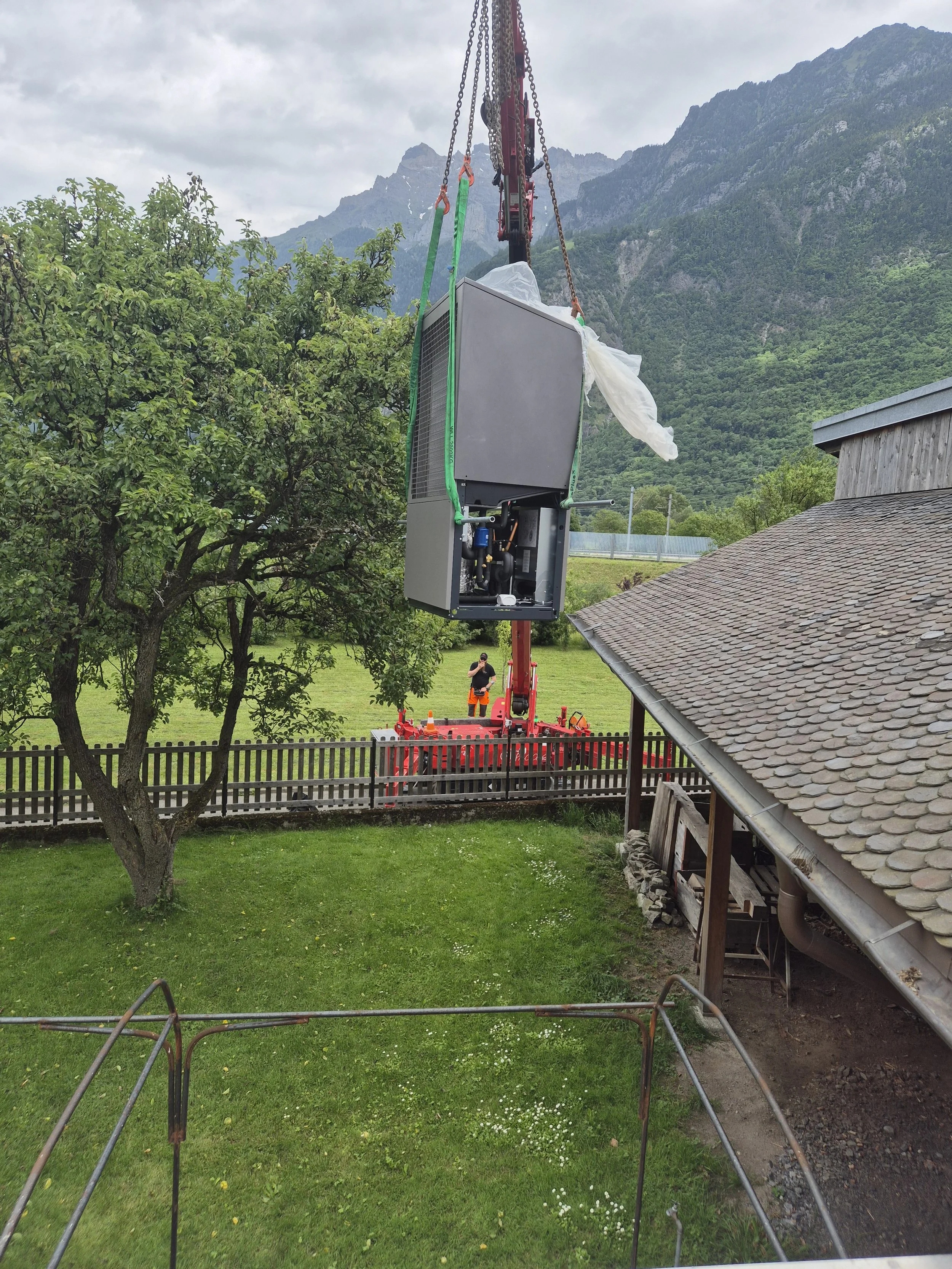 Un appareil de climatisation est suspendu par un grue dans un jardin avec un arbre, une maison et des montagnes en arrière-plan.