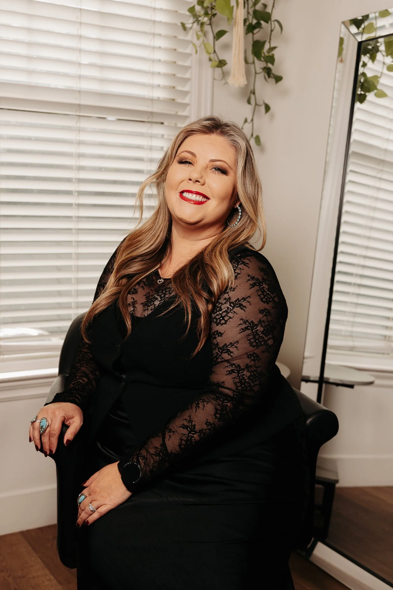 A smiling woman with long wavy blonde hair, wearing a black lace top with sheer floral detailing, sitting in a black chair next to a window with white blinds, a mirror, and a hanging plant in the background.