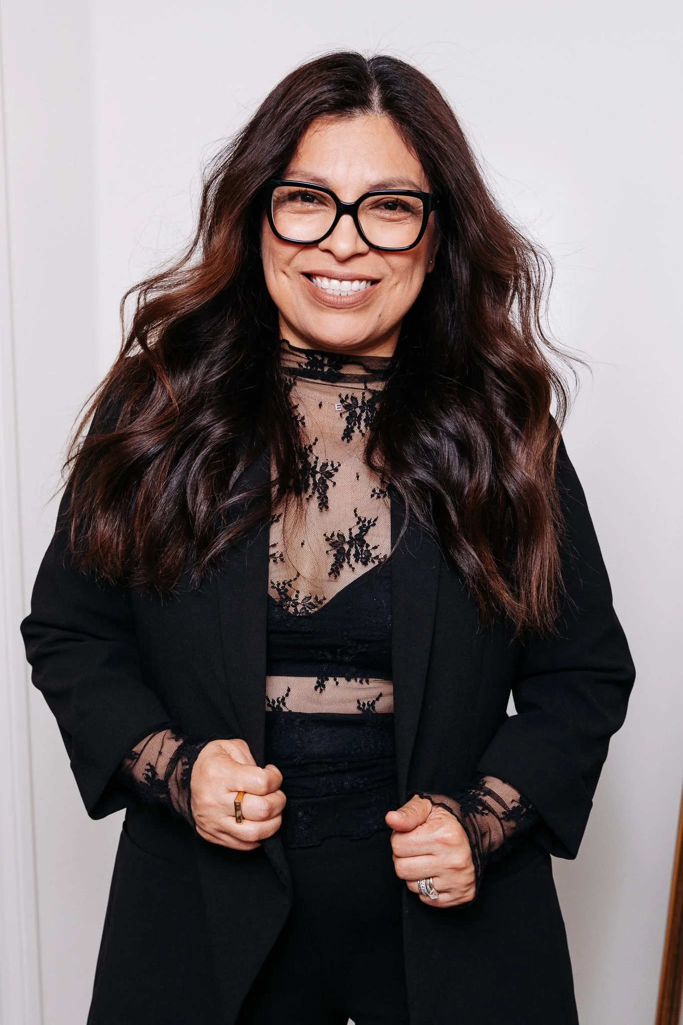 A woman with long dark brown hair, large black glasses, and a wide smile, wearing a black blazer over a sheer lace top, standing against a plain white background.