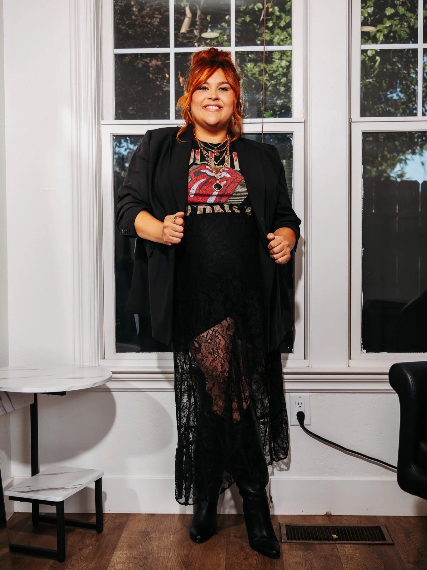A woman with red hair, dressed in black, standing in front of a window with green trees outside, indoors at night.