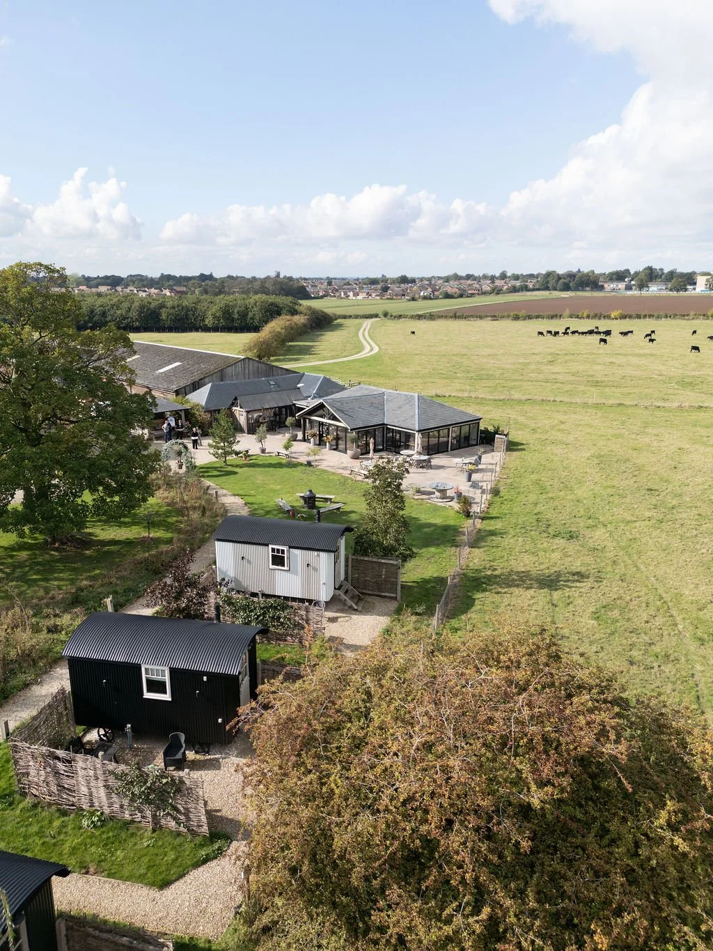 Aerial view of a rural property with modern black and white sheds, a building with a patio, and a large open field with grazing cows. There are trees, gravel pathways, and a distant village under a partly cloudy sky.