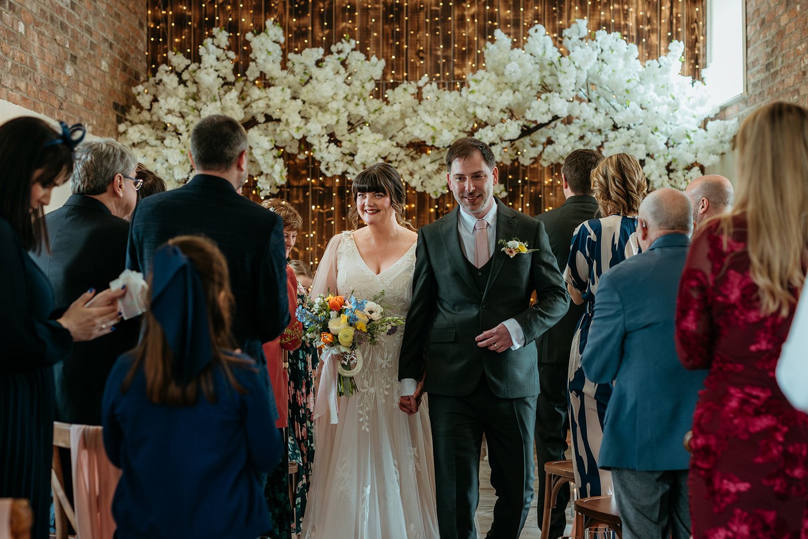 A bride and groom walking hand-in-hand down the aisle during their wedding ceremony in a decorated venue with floral arrangements and string lights.