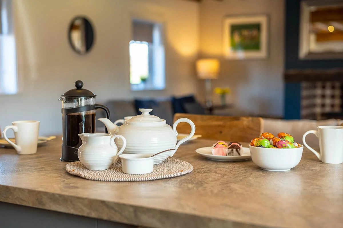 Tea set with a teapot, cream pitcher, sugar bowl, a French press, and bowls of colorful candies and cupcakes on a kitchen island.