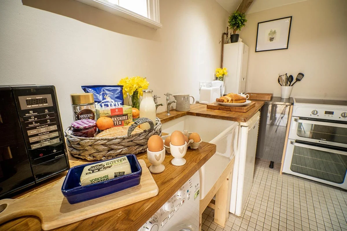 Kitchen counter with eggs in egg cups, a basket of groceries including eggs, tea, and coffee, a loaf of bread on a wooden cutting board, flowers in vases, and kitchen appliances.
