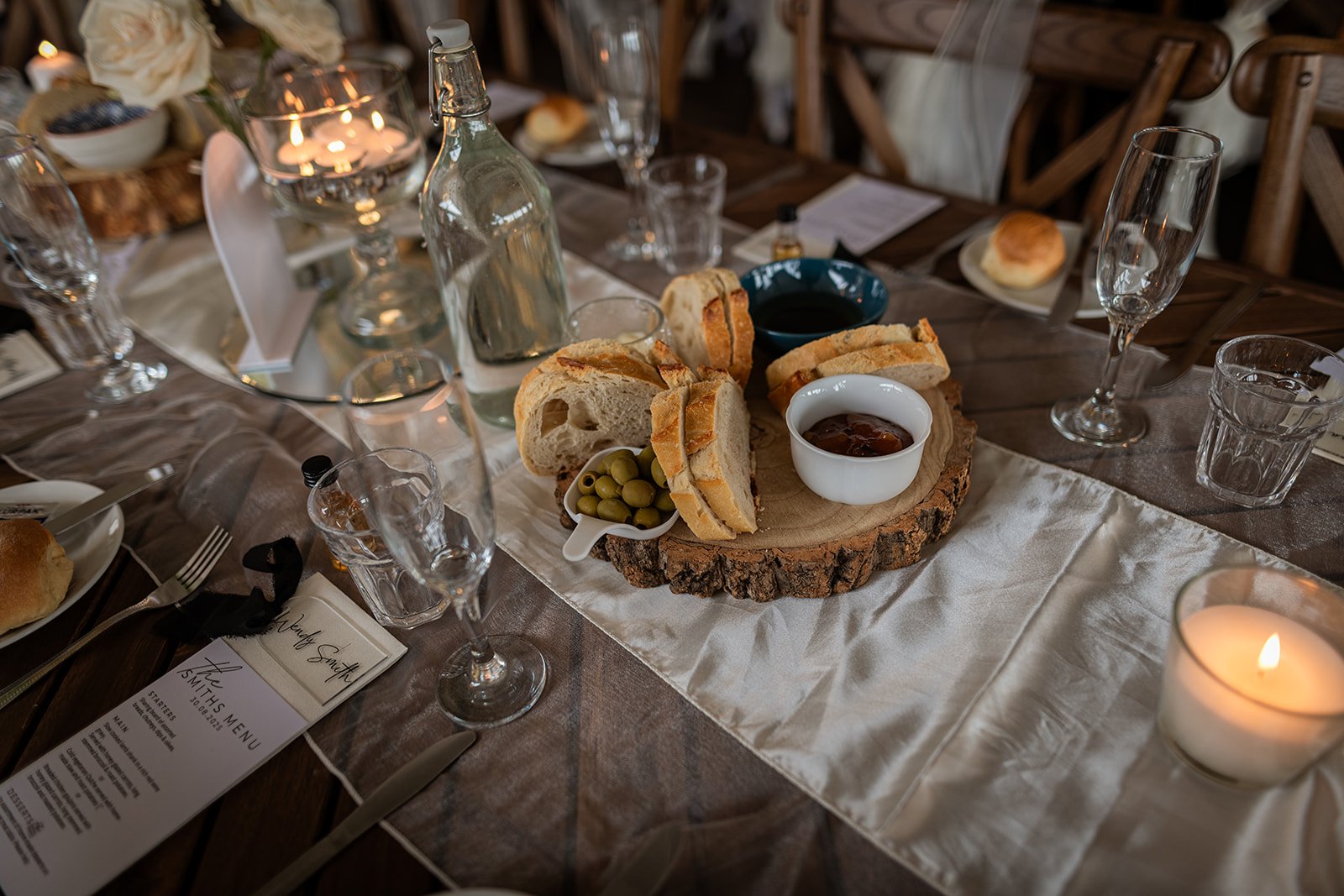 A rustic dining table set for a meal with bread, green olives, and condiments on a wooden and fabric table runner, surrounded by empty wine glasses, candles, and place cards.