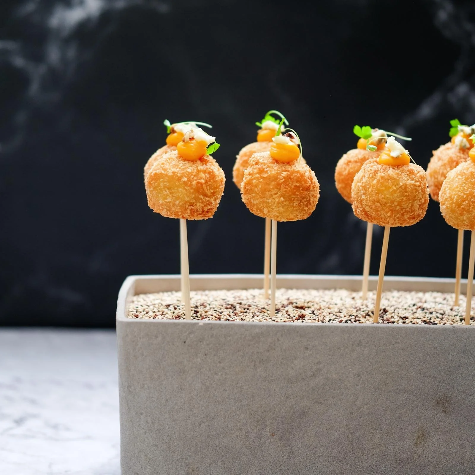 Appetizers on skewers with breaded coating, topped with small dollops of sauce, microgreens, and tiny edible flowers, arranged in a row in a rectangular container filled with sand or small beads.