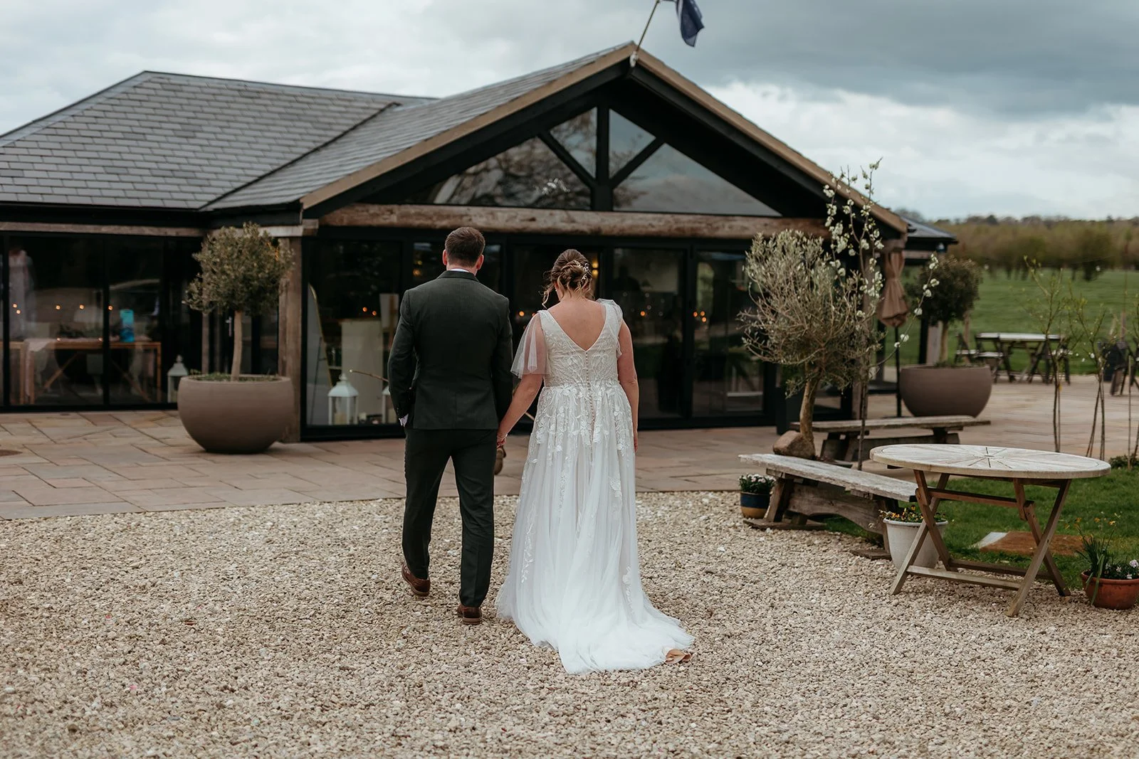 A bride and groom walking hand in hand outside a rustic wedding venue with glass doors, potted plants, and a gravel pathway, under an overcast sky.