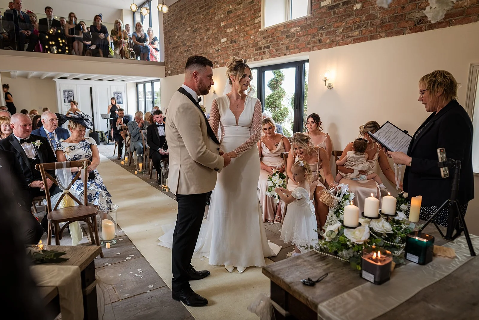 Bride and groom holding hands during wedding ceremony in a bright, rustic indoor venue with guests seated and standing, officiant reading vows, candles and floral decorations on table.