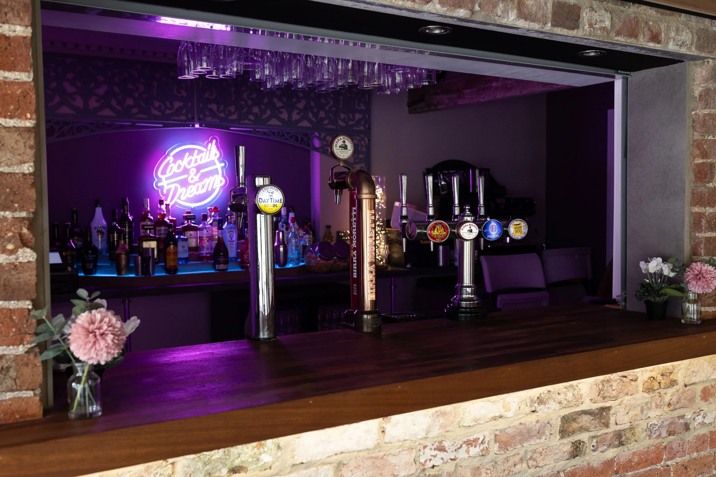 Bar area with bottles, taps, and glasses, illuminated by purple neon sign that reads 'Cocktails & Dreams,' with brick walls and floral decorations.