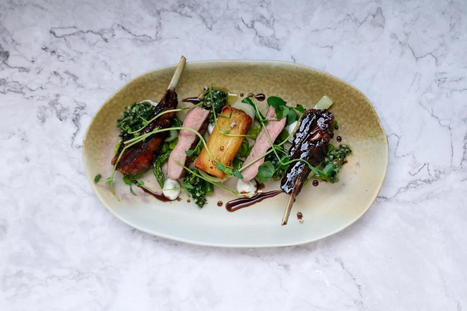 Gourmet dish on an oval plate with grilled meat, vegetables, microgreens, and a dark sauce, served on a marble surface.