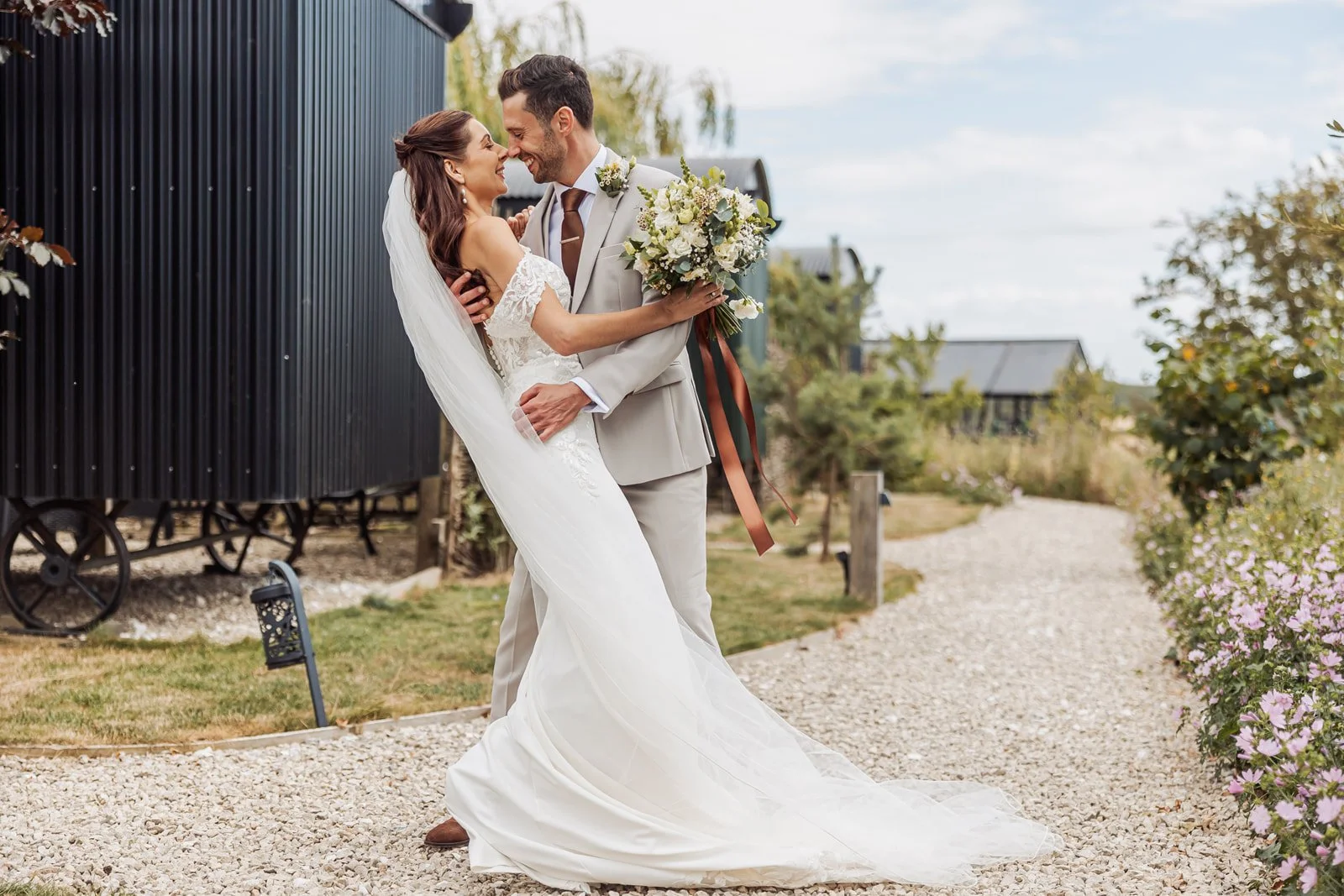 A bride and groom smiling and embracing outdoors on a wedding day, with the bride holding a bouquet, standing on a gravel path surrounded by greenery and flowers.