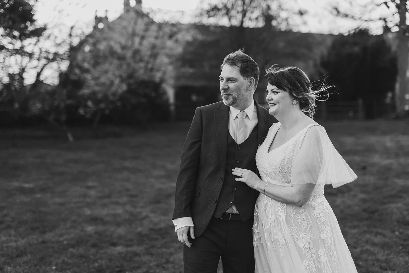 A black-and-white photo of a couple in wedding attire standing outdoors on a grassy area with trees in the background, smiling and looking away from the camera.