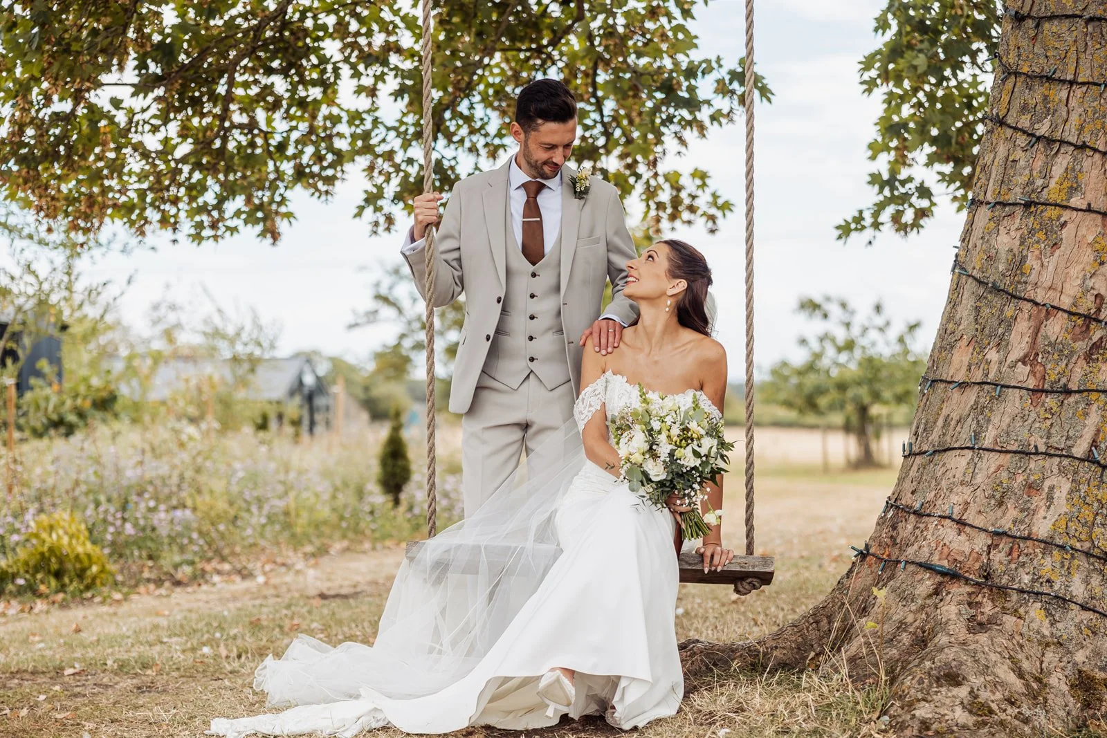 A bride in a white wedding dress sitting on a swing under a tree, holding a bouquet, with a groom in a light gray suit standing beside her, in an outdoor setting.