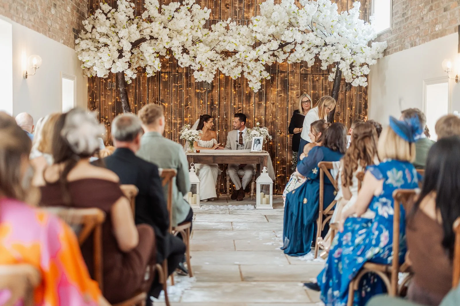 A wedding ceremony with a bride and groom sitting at a table, surrounded by guests seated on wooden chairs, with decorative white flowers and fairy lights on a wooden backdrop.