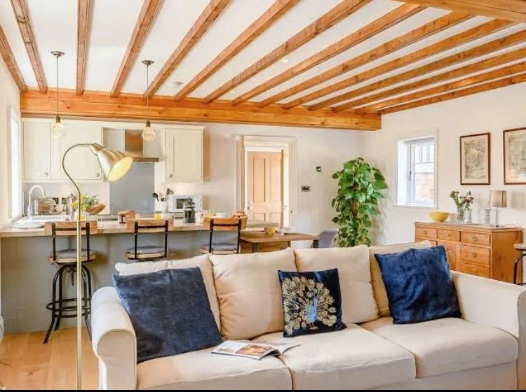 Living room and kitchen in a cozy home with a wooden ceiling, beige sofa with decorative pillows, a tall floor lamp, and a wooden sideboard. The kitchen has a counter with bar stools, white cabinets, and open shelving, with a door leading to another 