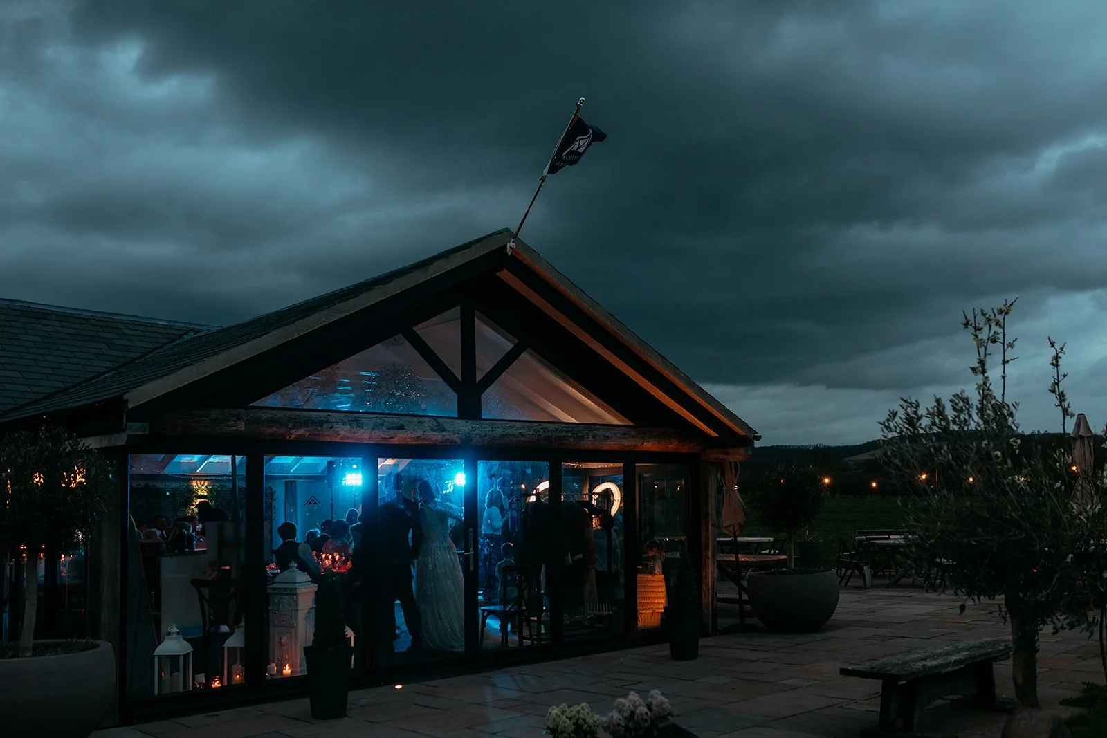 Nighttime outdoor wedding reception inside a glass-enclosed barn with dark stormy skies overhead. Guests are dancing and celebrating, illuminated by blue and orange lights.