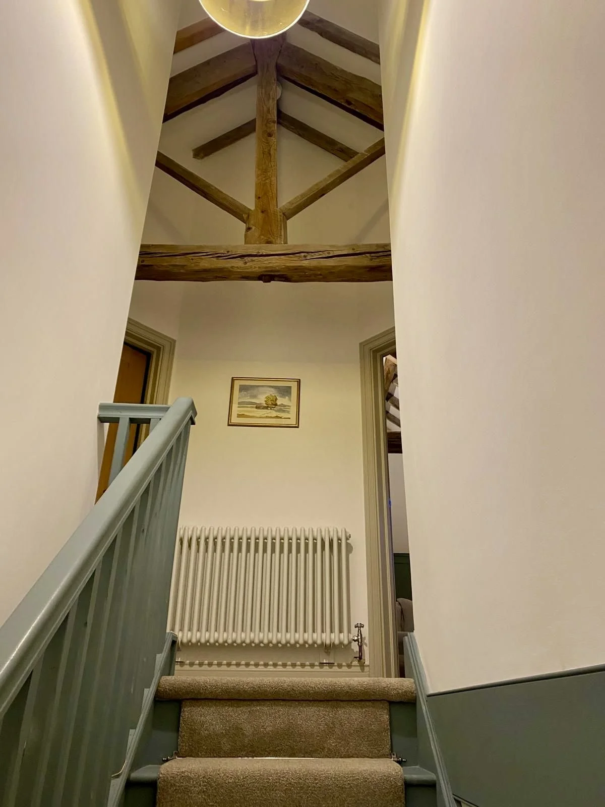 Interior view of a staircase leading up to a landing with a wooden beam ceiling, a fitted picture frame of landscape, a radiator, and beige carpeted stairs.