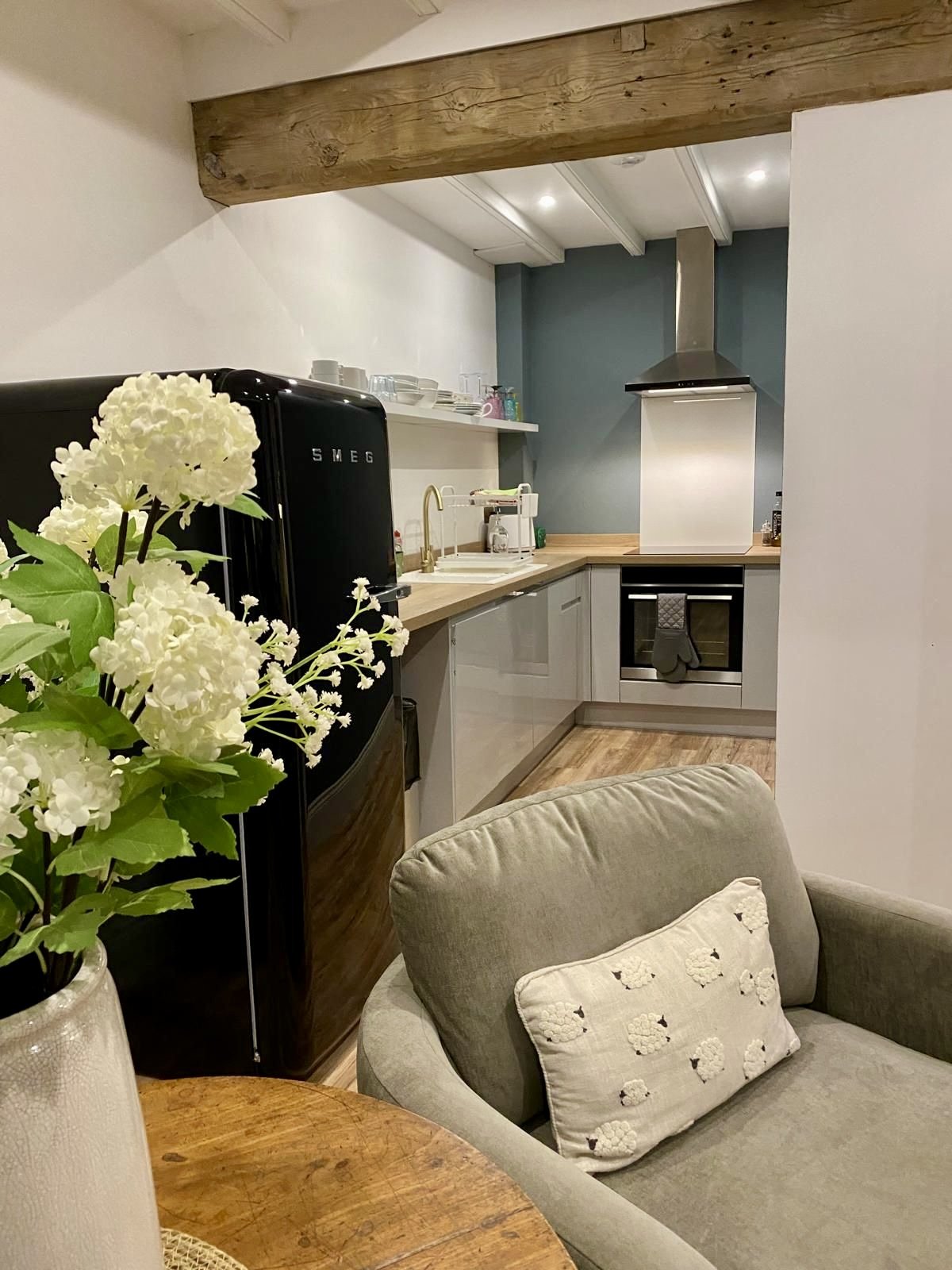 A cozy kitchen with a black SMEG refrigerator, a wooden countertop, white cabinets, a stove with a tea towel hanging, and shelves with dishes and containers. There is a beige armchair with a decorative pillow in the foreground and a vase with white f