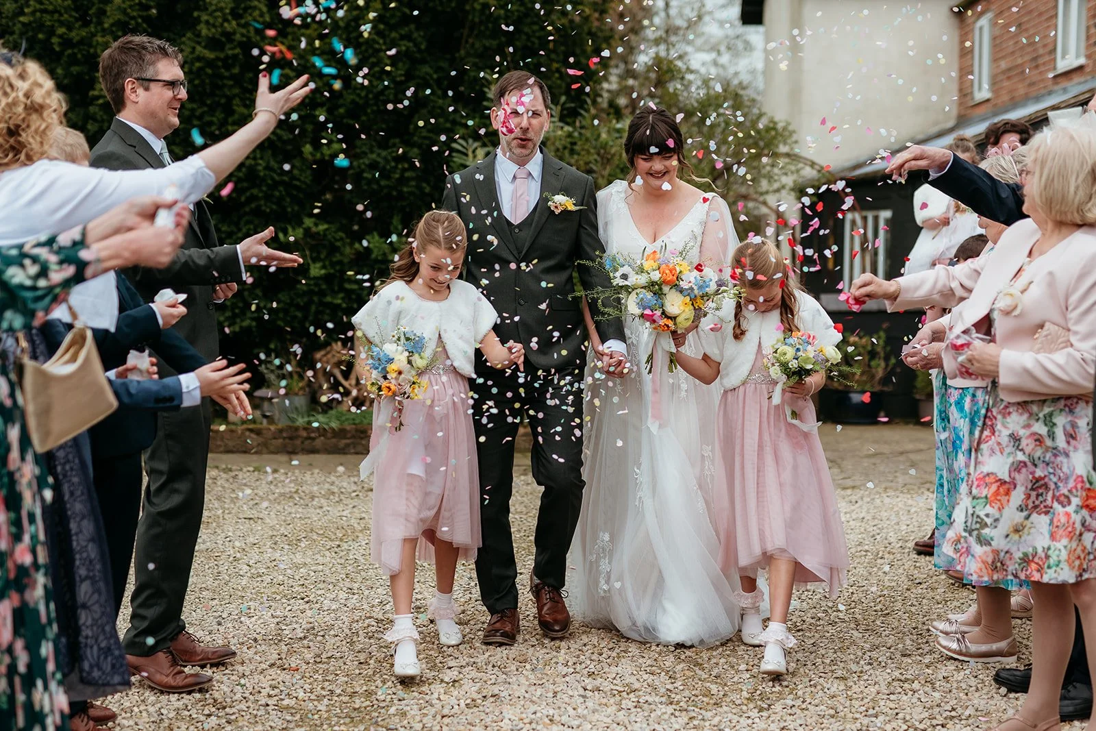 A bride and groom walking outdoors on their wedding day, surrounded by guests throwing colorful confetti. The bride is holding a bouquet and smiling, while the groom is holding her hand. There are two flower girls holding bouquets and walking alongsi