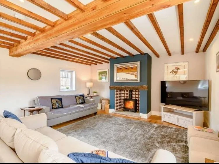 Living room with a wood-beamed ceiling, a fireplace with a mirror above it, a flat-screen TV on a white stand, a large beige sofa, a gray rug, and artwork on the walls.