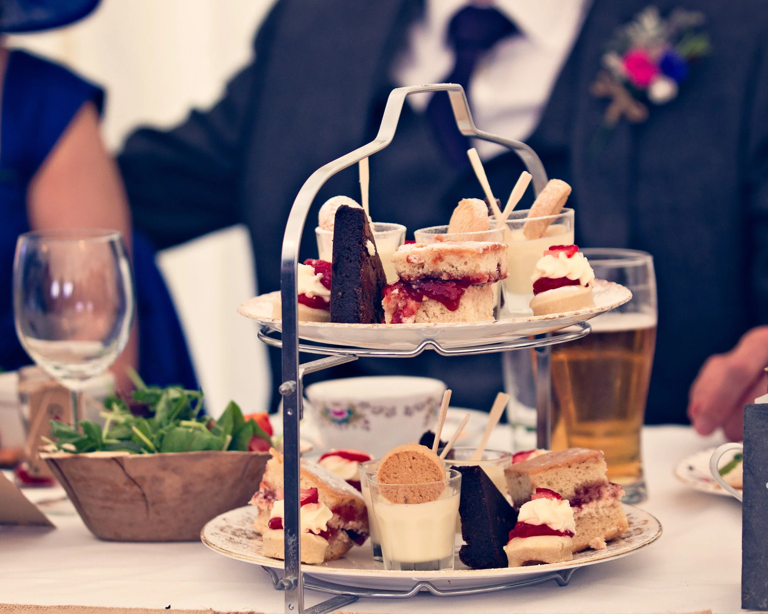 Two-tiered serving tray filled with assorted desserts, including slices of cake, cookies, and cream-filled pastries, with a bowl of salad and glasses of drinks nearby, indicating a buffet setting for a celebration or gathering.