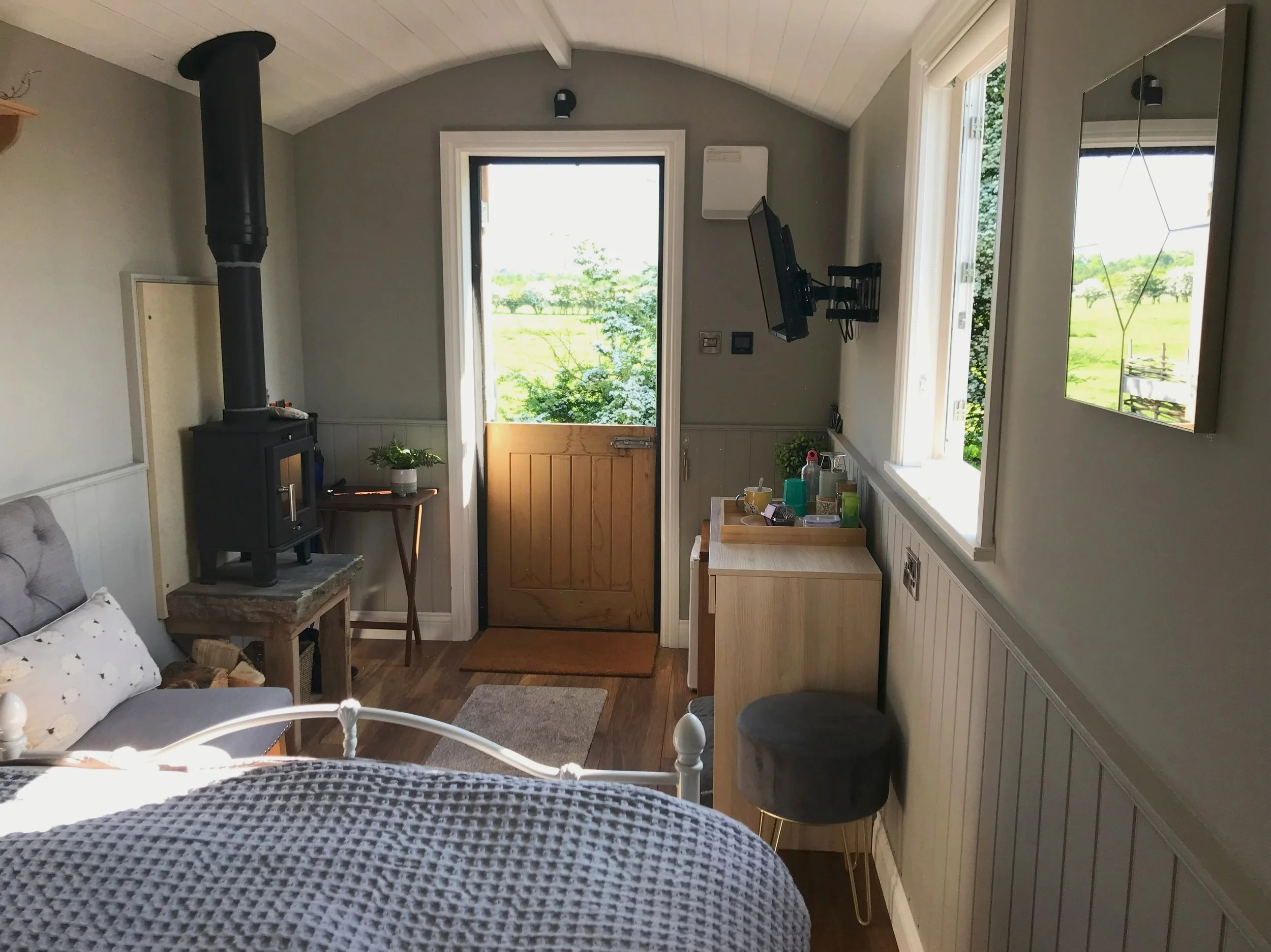 Cozy bedroom with a white iron bed, gray pillow, wood stove in the corner, small table with a plant, wood door with window leading to a green landscape, and two windows reflecting the view.