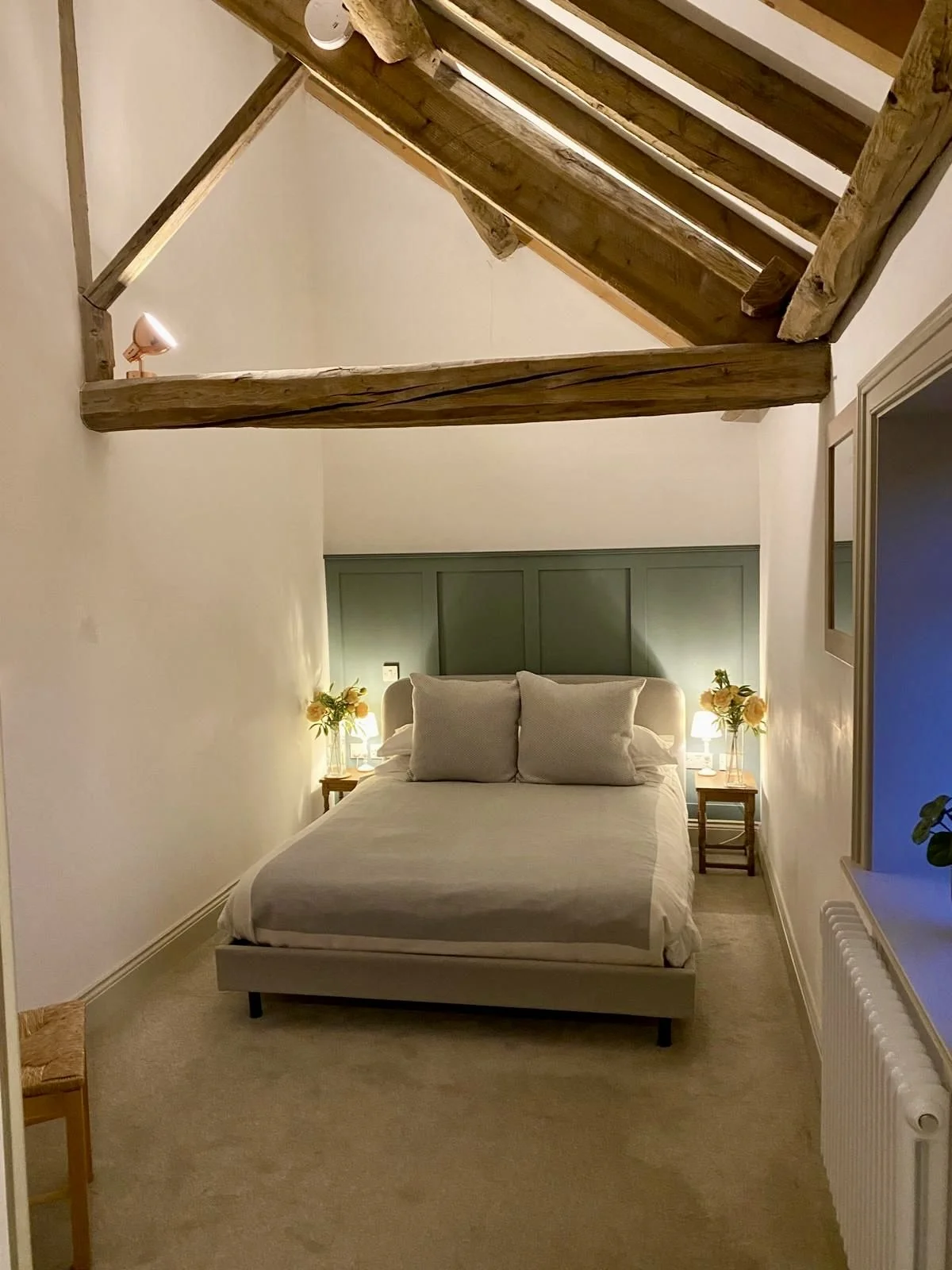 A cozy bedroom with a bed against a green headboard, two bedside tables with lamps and yellow flowers, beige carpet, white walls, and an exposed wooden beam ceiling.