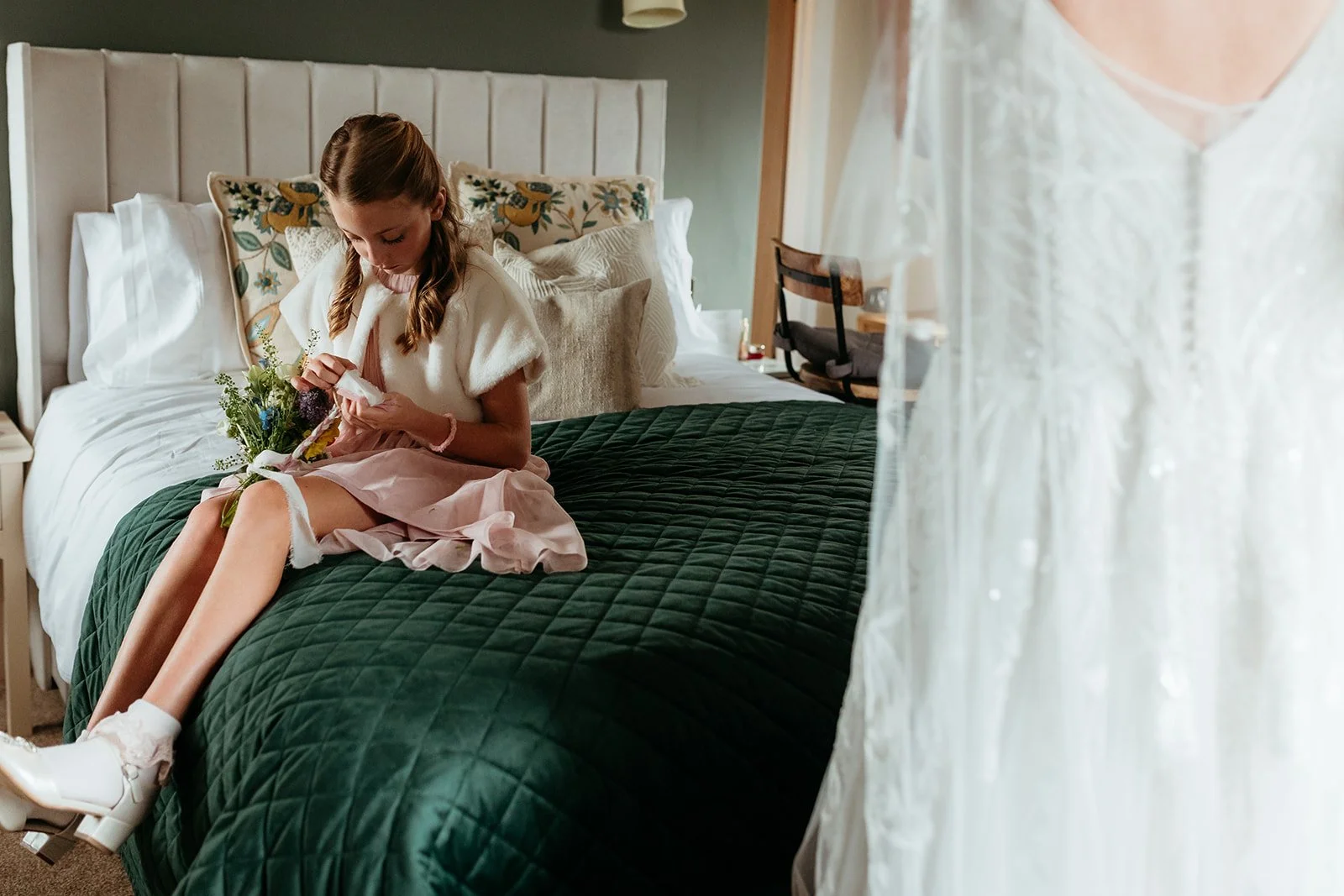 A young girl in a pink dress and white shoes sitting on a green quilted bed, looking at a small item in her hands with a bouquet of flowers on her lap. The bed has a white upholstered headboard and several decorative pillows. A curtain is partially hanging in the foreground.