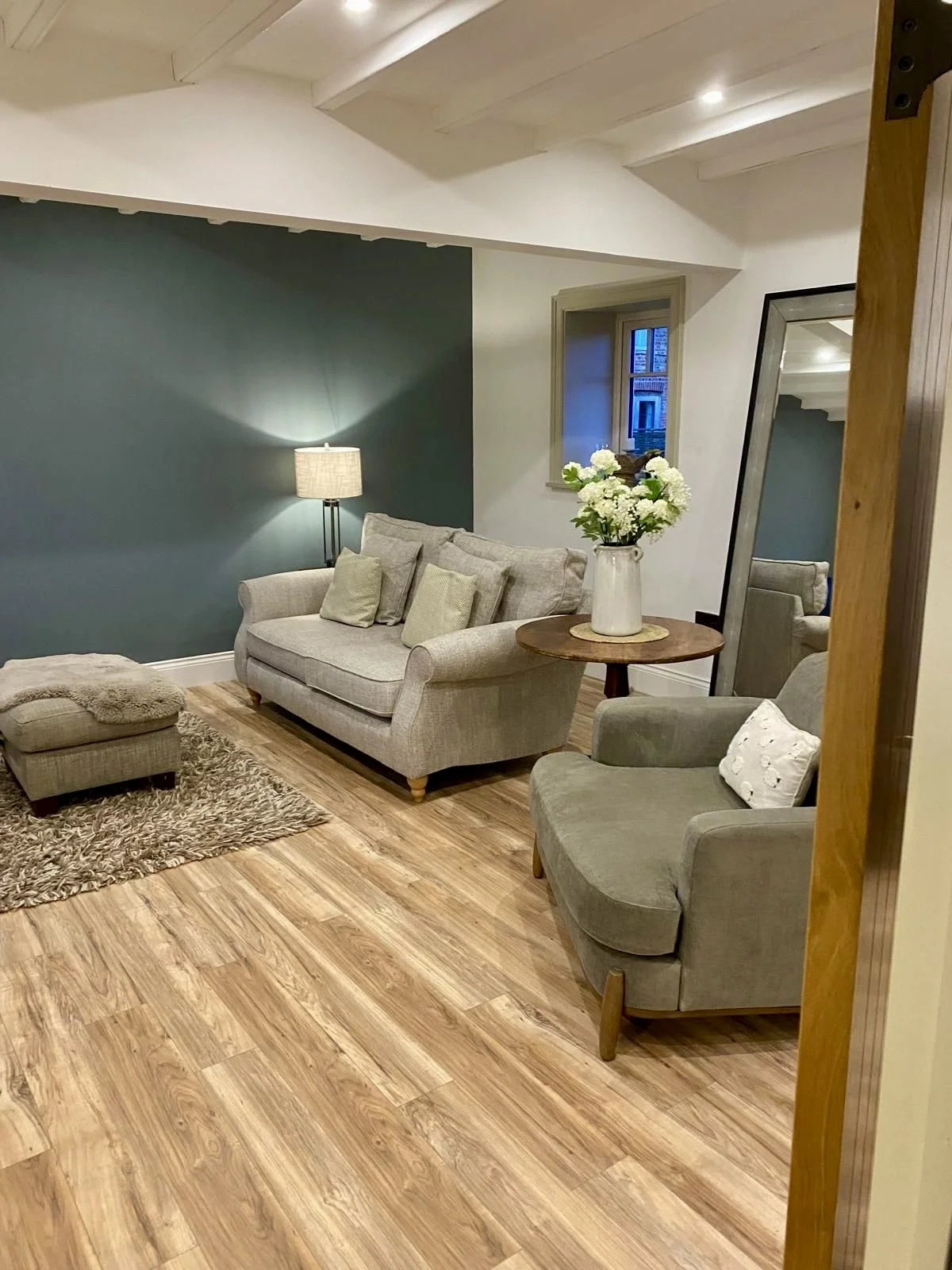 Cozy living room with beige furniture, a blue accent wall, a lamp, a wooden side table with a vase of white flowers, a large mirror, and hardwood floors.