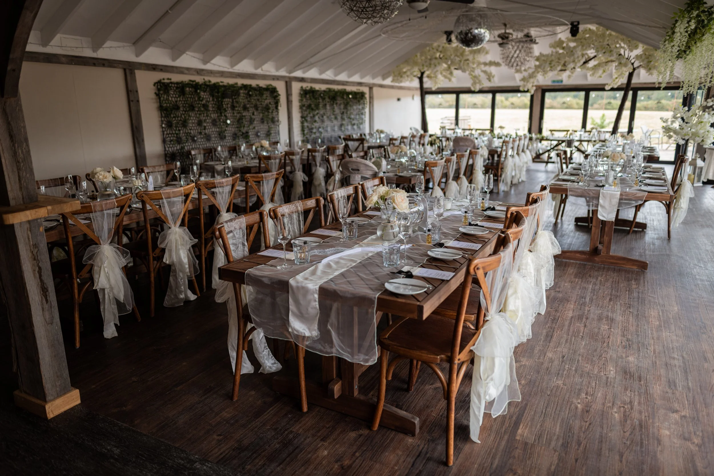 Elegant wedding reception setup with long wooden tables, white floral centerpieces, and cream-colored chair sashes, inside a decorated rustic hall with large windows and hanging floral arrangements.