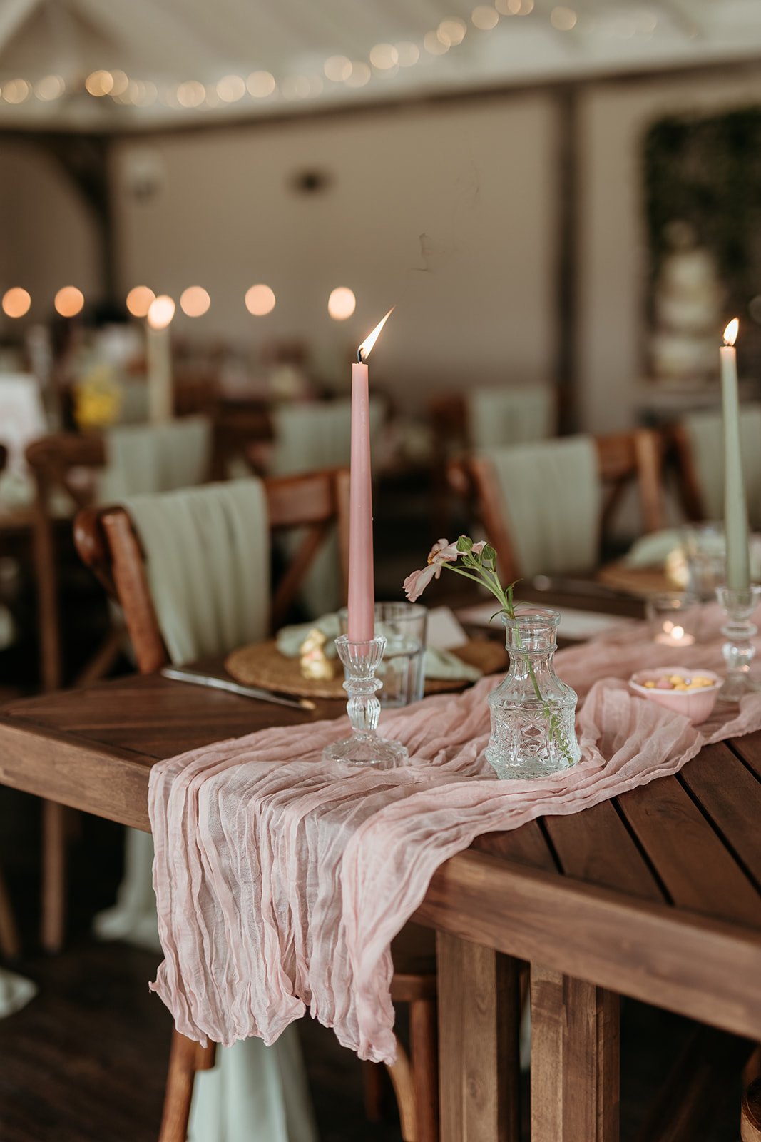 A decorated dining table with pink and green candles, small vases with flowers, and a pink table runner in a cozy, dimly lit setting with string lights overhead.