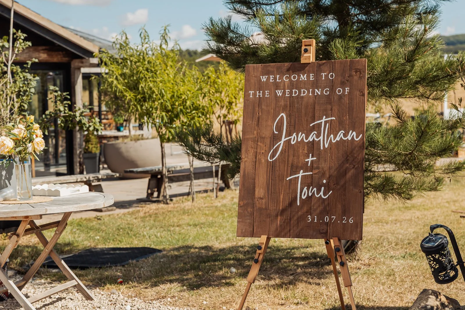 Wooden wedding sign welcoming guests to the wedding of Jonathan and Toni, with the date 31.07.26, outdoors with greenery, flowers, and a rustic setting.