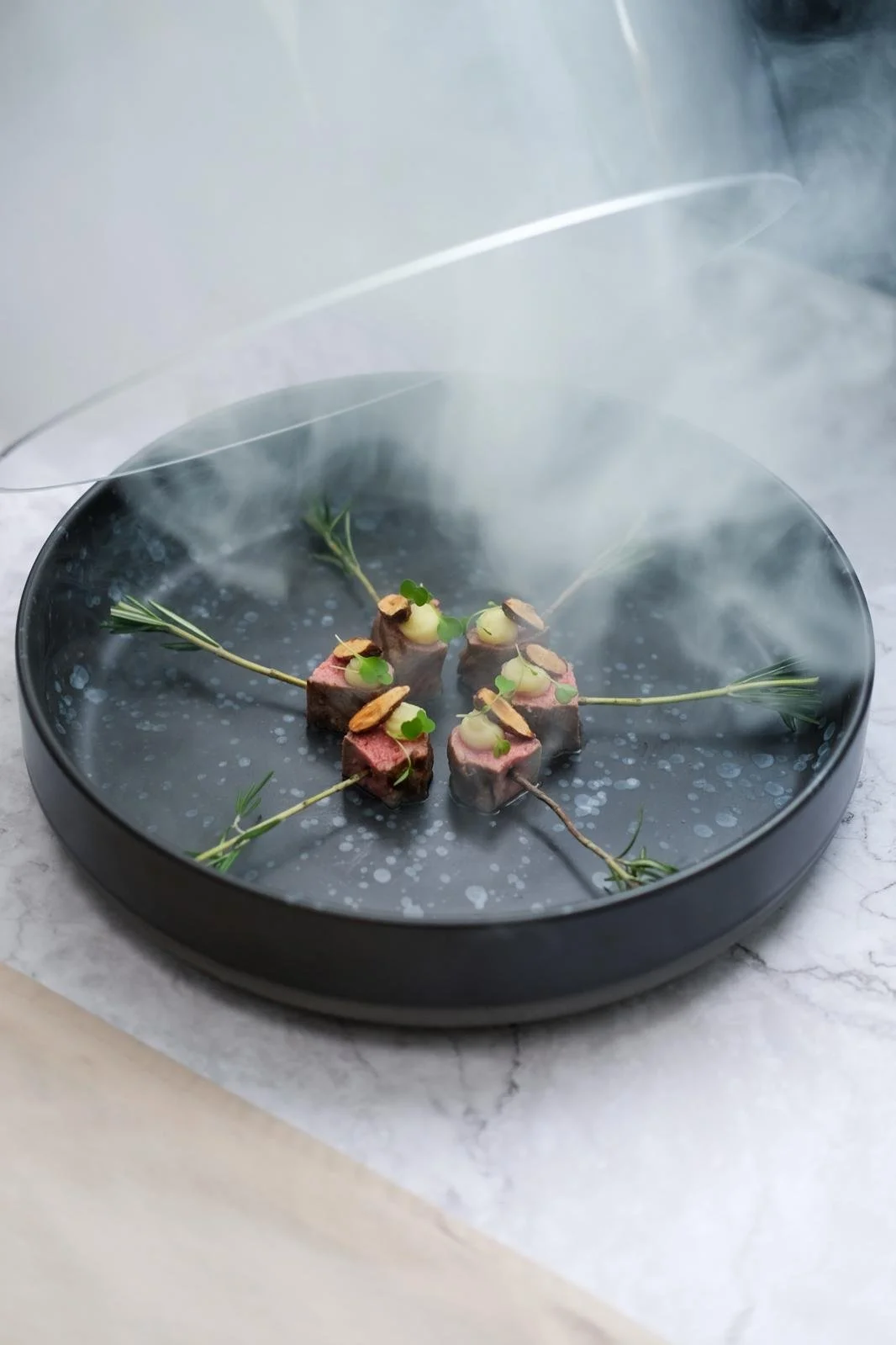 Modern dish presentation with small pieces of beef, garnished with herbs, thin vegetables, and edible flowers, served on a black round plate with visible smoke, on a marble surface.