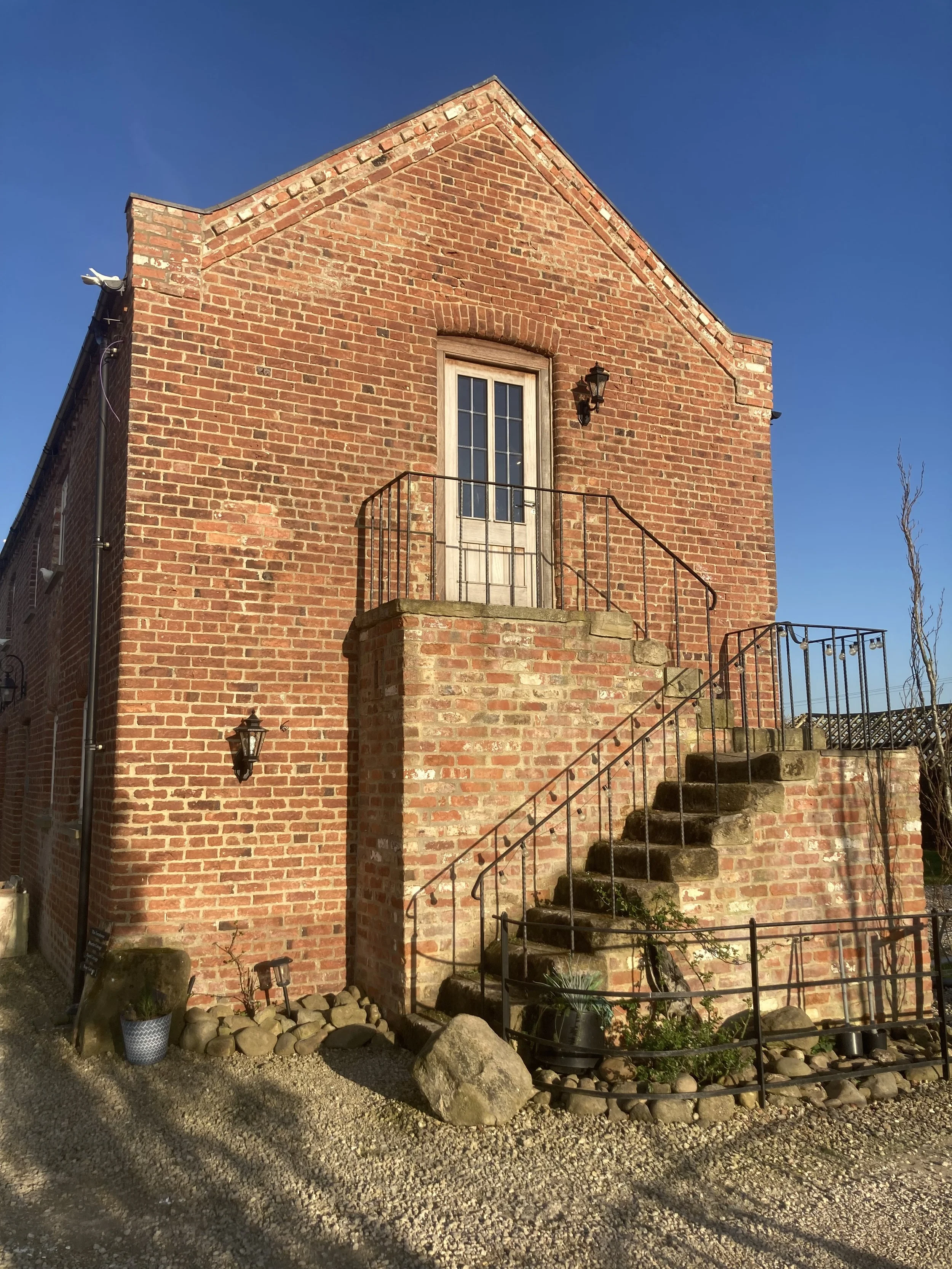 Front view of a two-story brick building with stairs leading to a door on the upper level, outdoor lantern lights, rocks and plants at the base, under a clear blue sky.