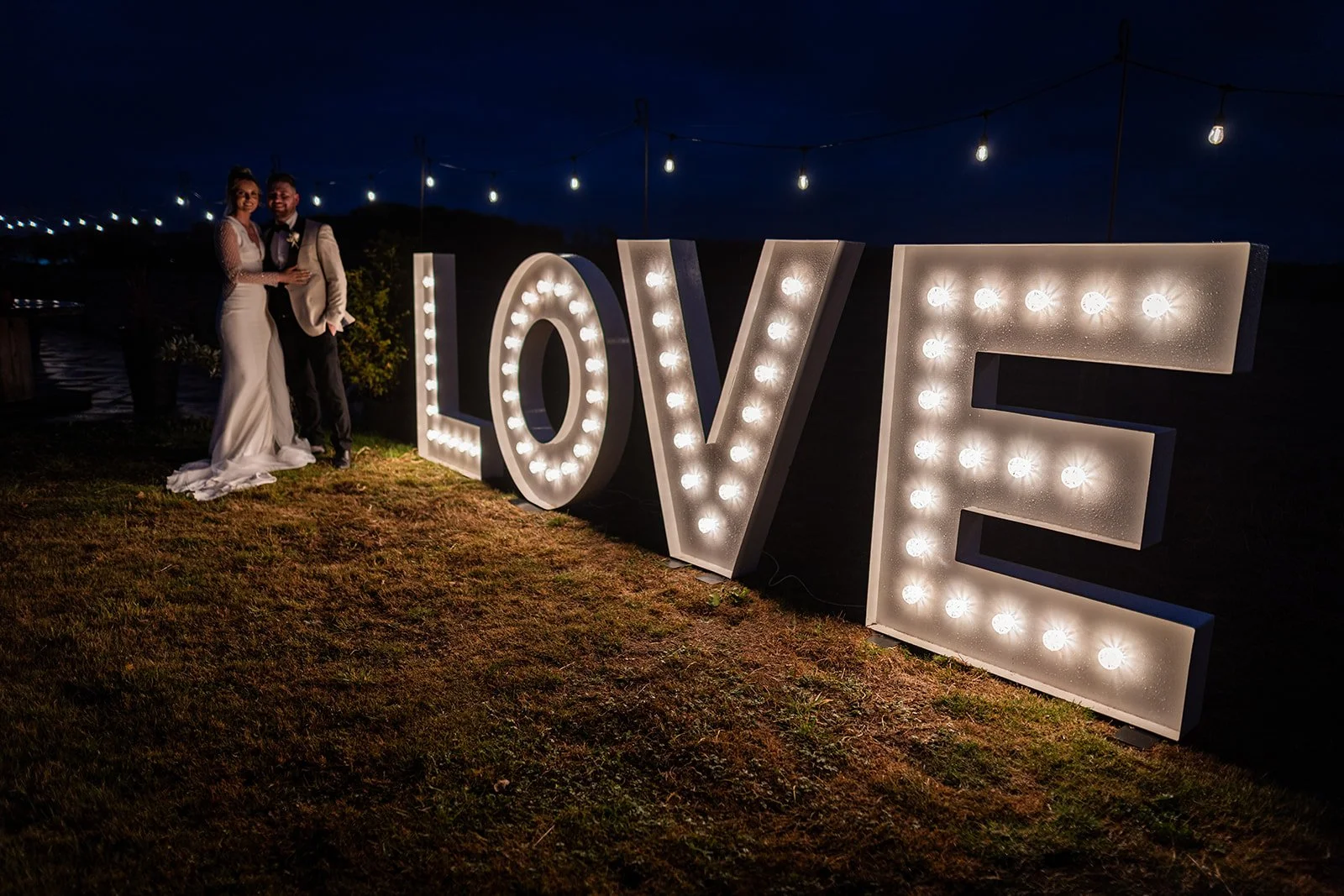 A couple in wedding attire stands beside illuminated large 'LOVE' sign outdoors at night.