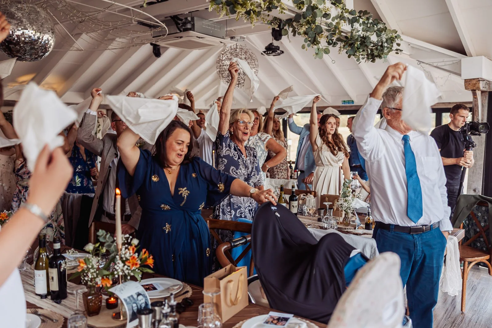 People at a celebration, dancing and waving napkins in a decorated indoor venue with tables, flowers, and bottles of wine.