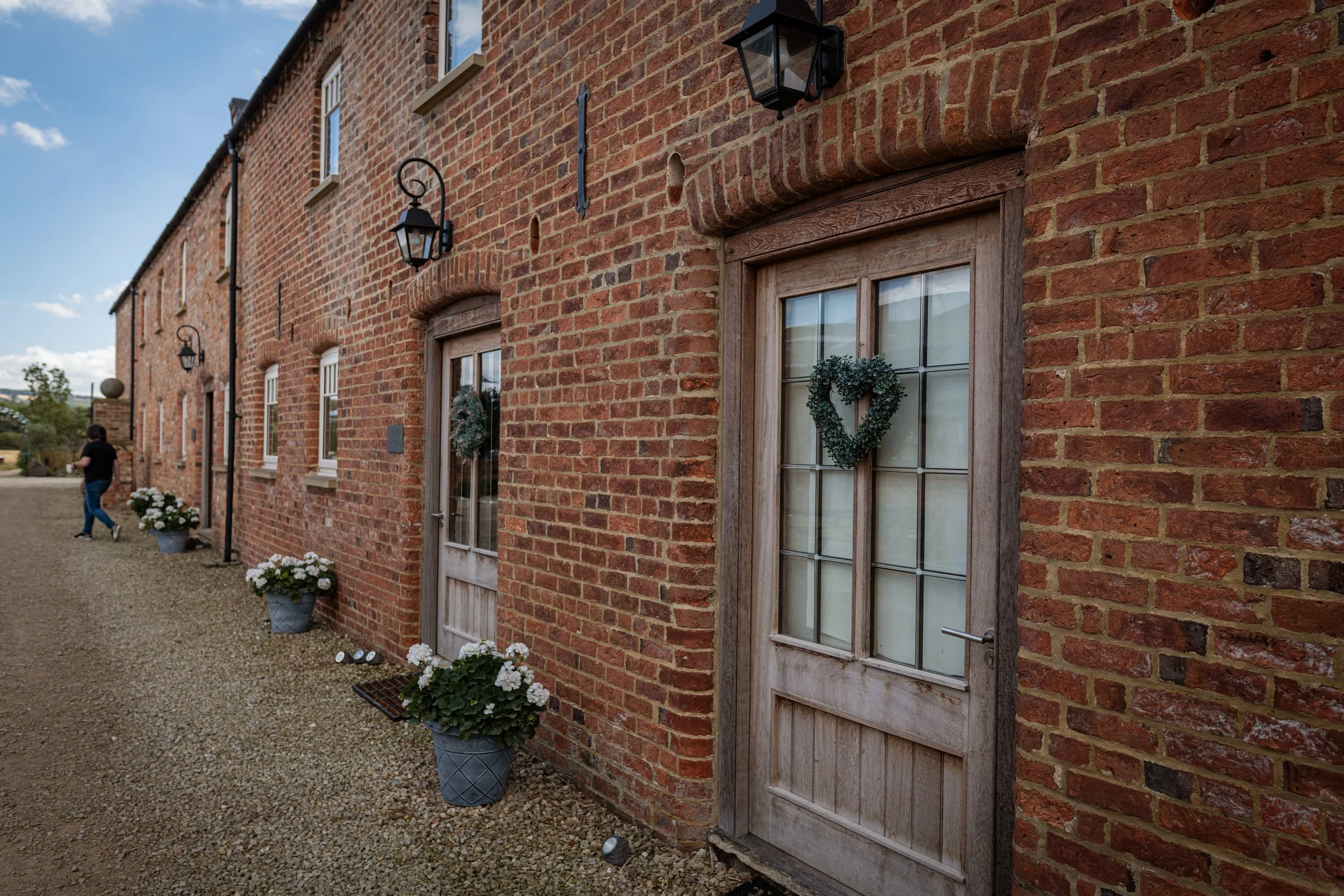 Brick building with wooden doors decorated with wreaths, potted white flowers outside, black lanterns, gravel pathway, person walking, cloudy sky.