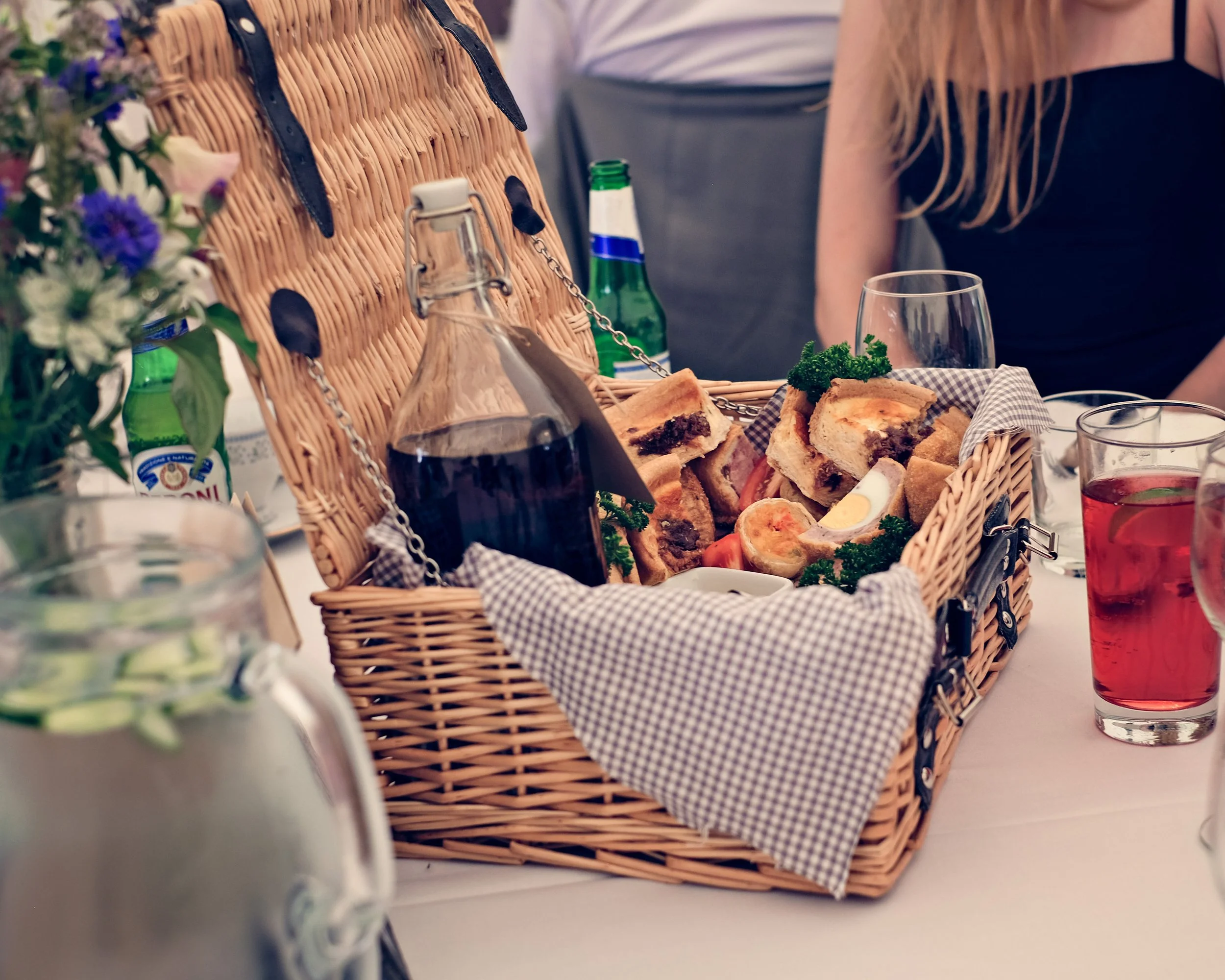 A wicker picnic basket filled with various sandwiches, a boiled egg, and garnished with parsley, placed on a table set for a meal.