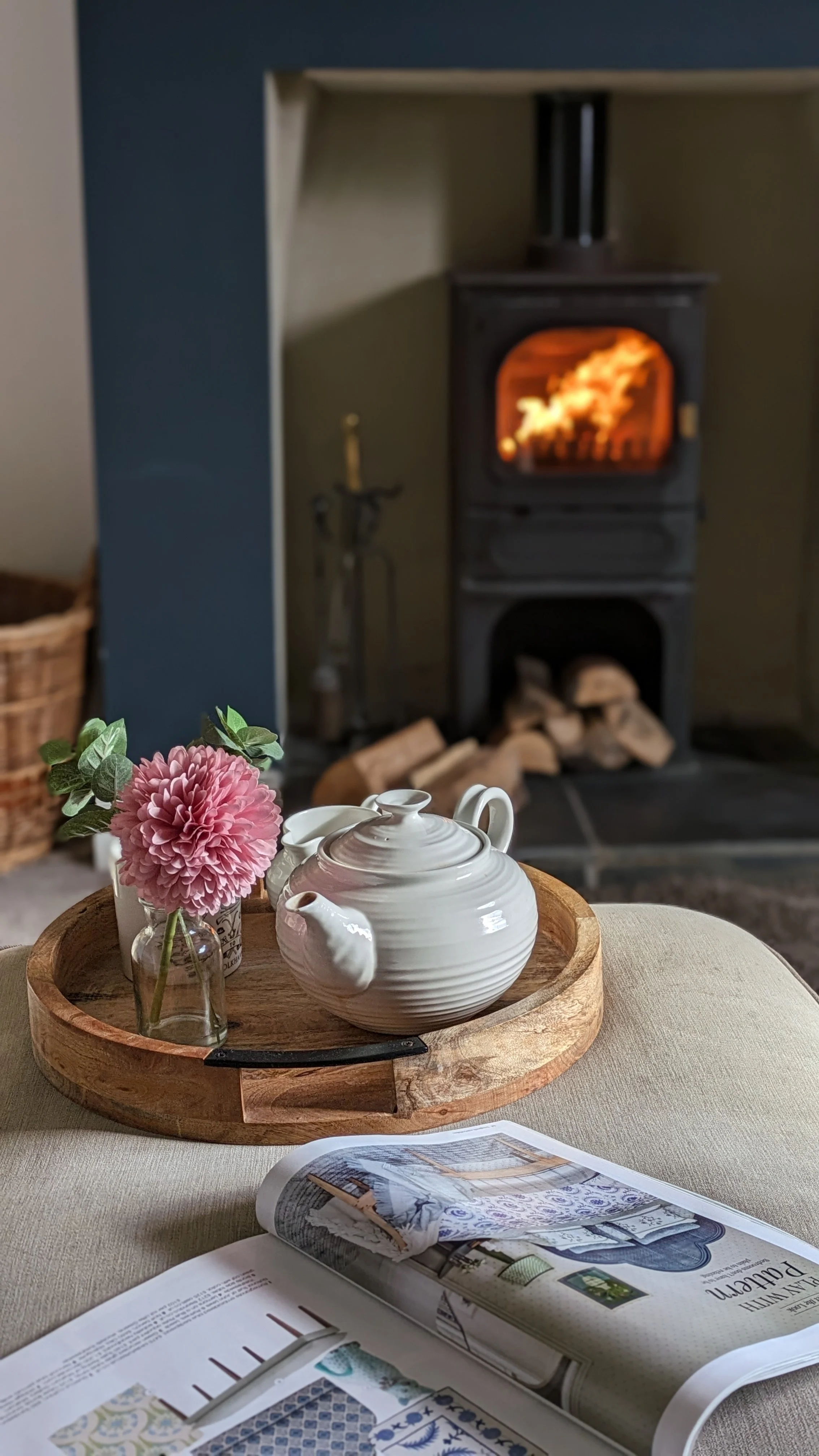 A cozy room with a wood stove with a fire burning inside, firewood stacked underneath, and fireplace tools nearby. In the foreground, a table holds a vase with a pink flower, a white teapot, and an open magazine.