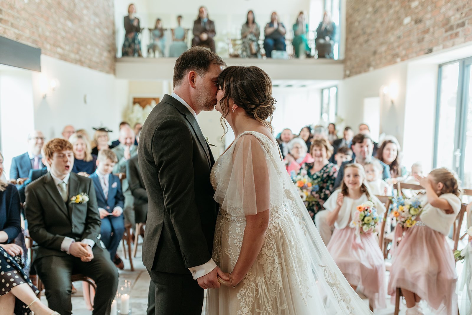 A bride and groom sharing a kiss during their wedding ceremony, with family and friends seated in a bright, rustic venue behind them.