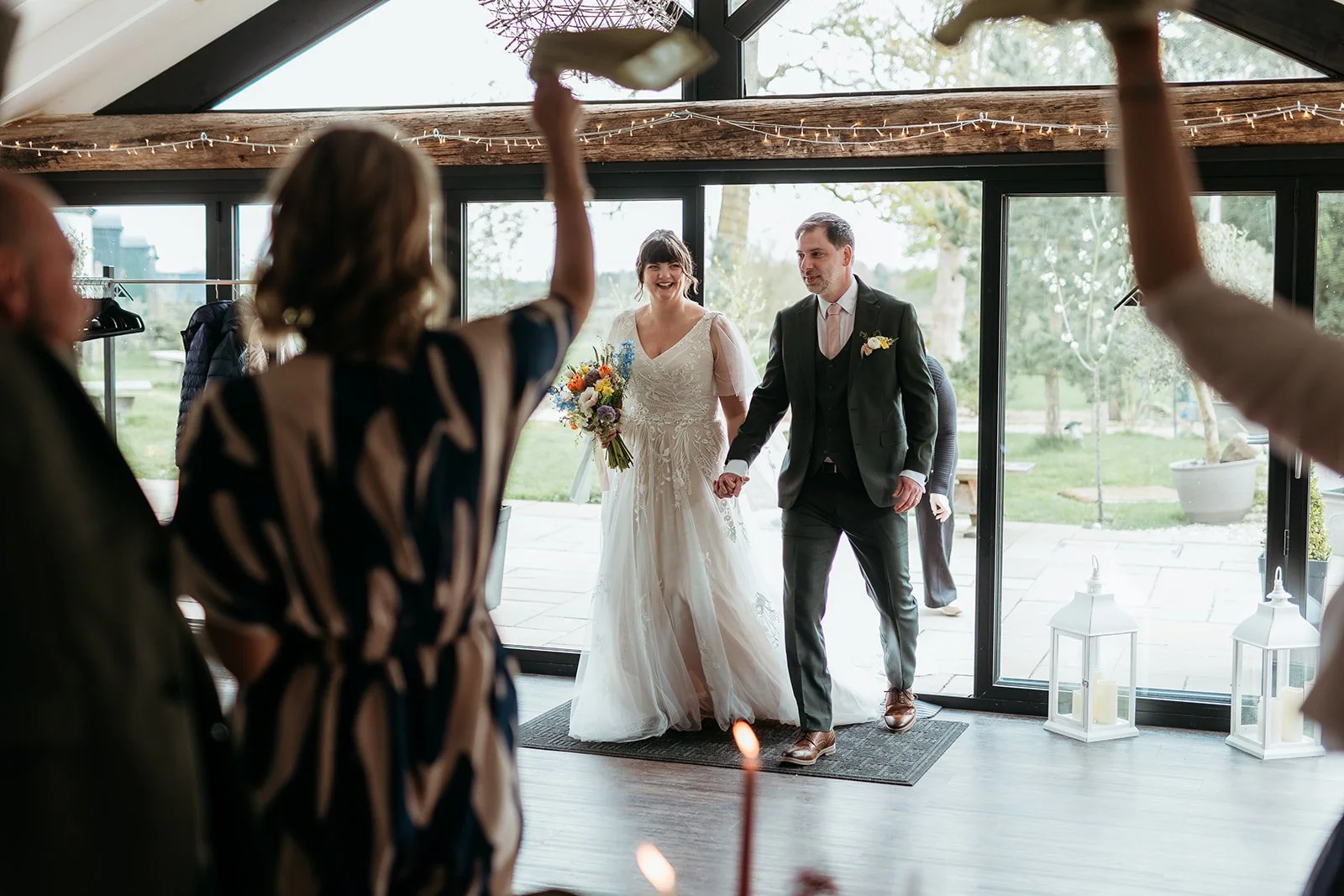 A bride and groom holding hands, entering a room during their wedding celebration, with guests in the foreground and candles in lanterns on the floor.