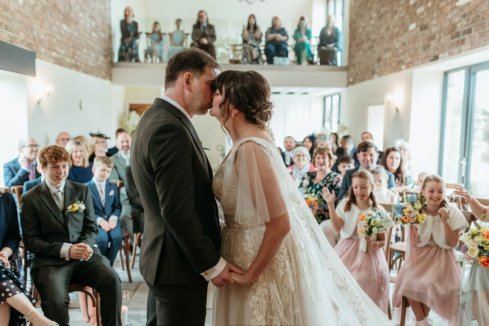 A bride and groom share a kiss during their wedding ceremony, with guests smiling and celebrating in the background.