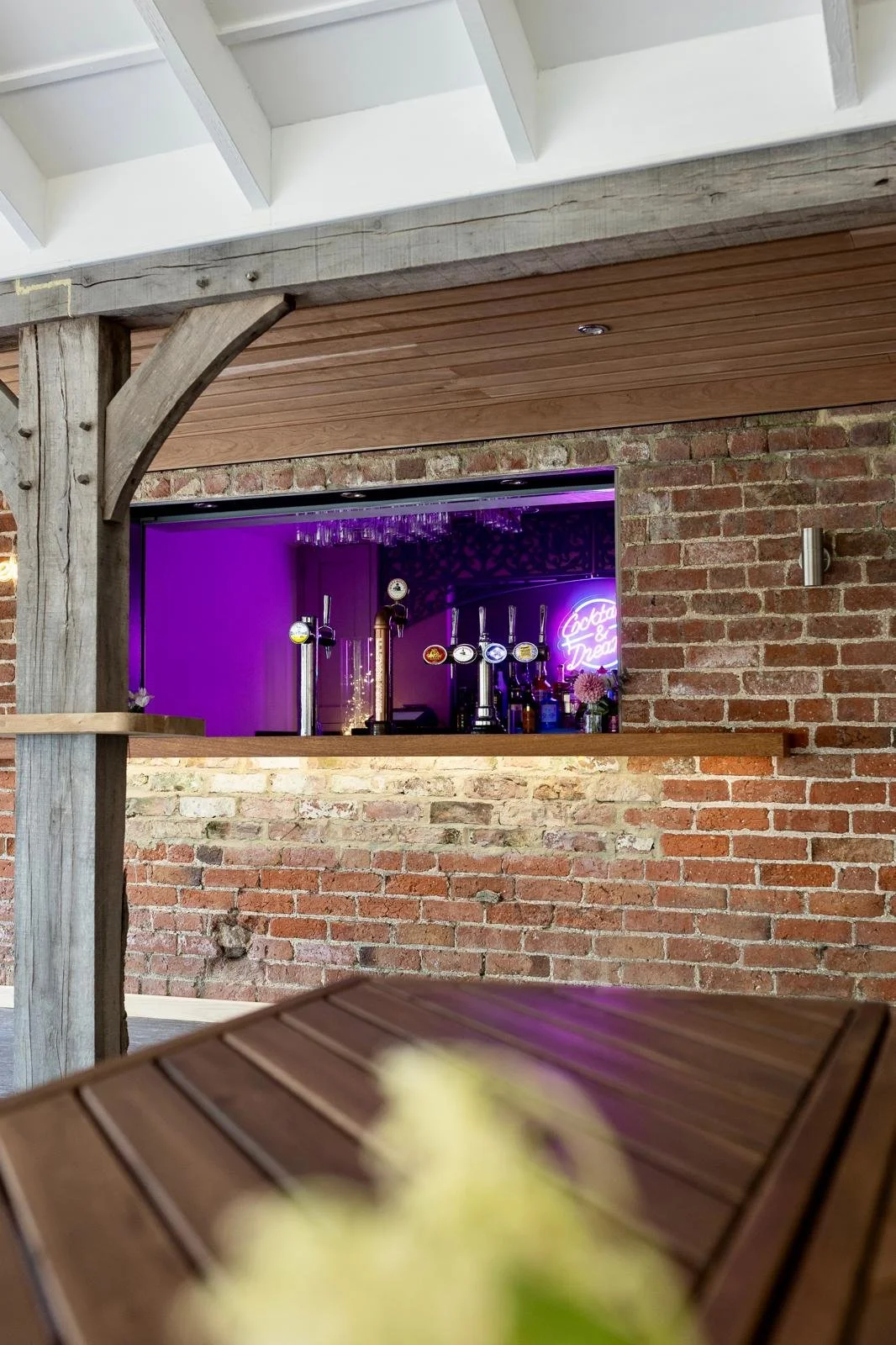 Bar area with 몇 taps, purple neon sign, flowers, and brick wall, viewed through a wooden table and a wooden beam.